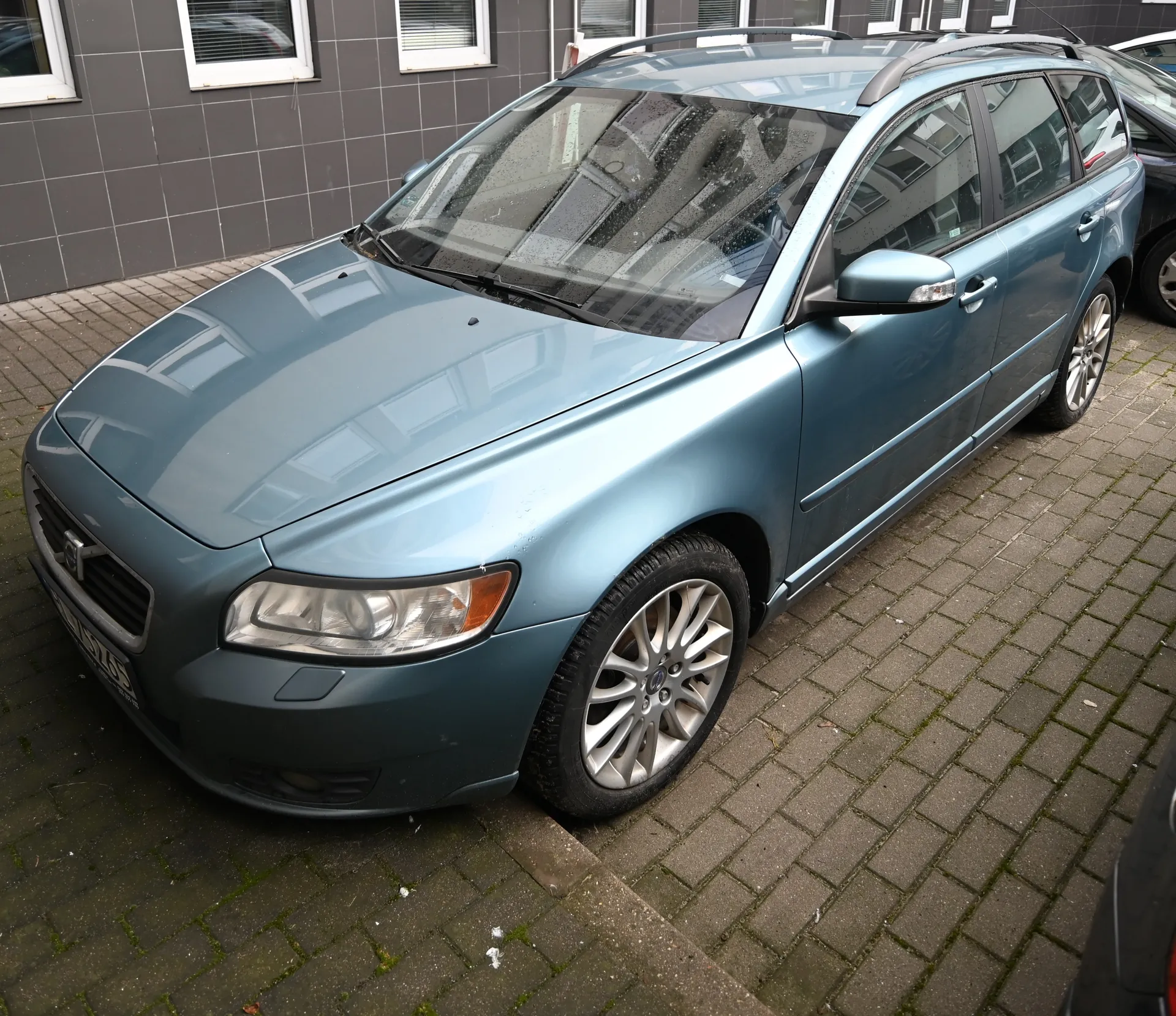 Niebieski samochód osobowy typu kombi marki Volvo zaparkowany na brukowanej powierzchni. Pojazd ma dachowe pręty, srebrne felgi i reflektory. W tle widoczny budynek z szarymi płytkami i białymi oknami. Na tablicy rejestracyjnej częściowo widoczny numer. Samochód znajduje się na chodniku z cegieł, a na powierzchni są ślady mchu.