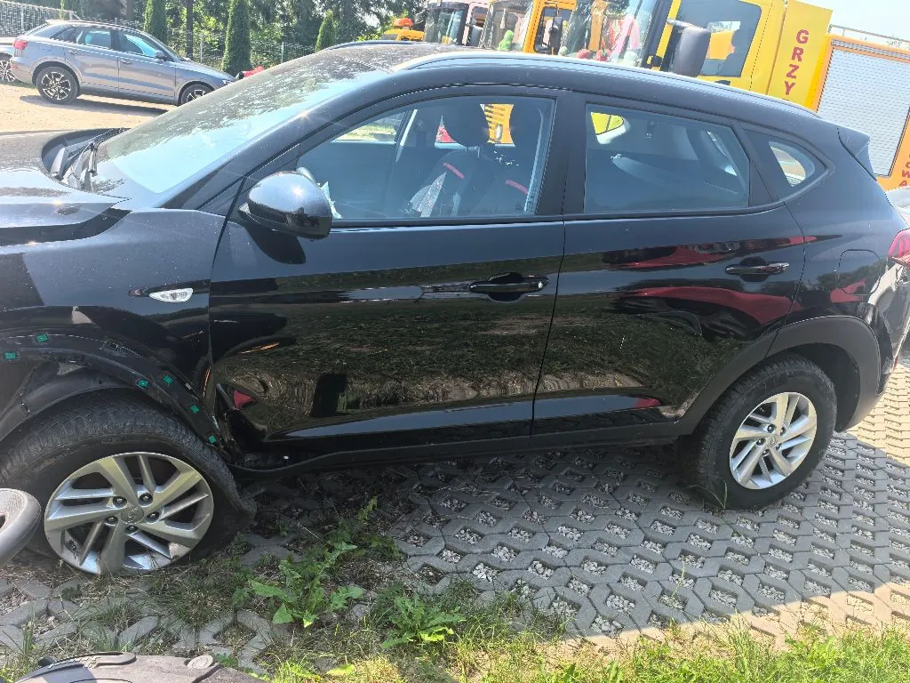 Czarny samochód typu crossover zaparkowany bokiem na szarym bruku z trawą wyrastającą w szczelinach. W tle widoczny jest inny, srebrny samochód oraz fragmenty żółtego sprzętu budowlanego z napisem 'GRNY'.