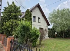 Jednopiętrowy dom z dachem dwuspadowym, o jasnej elewacji, z dwoma oknami i drzwiami wejściowymi. Dom otoczony jest zielonymi krzewami i drzewami. W pierwszym planie widoczny ceglany mur z czarną metalową bramą. Tło stanowi trawnik i niebo z chmurami. Brak czytelnego tekstu.