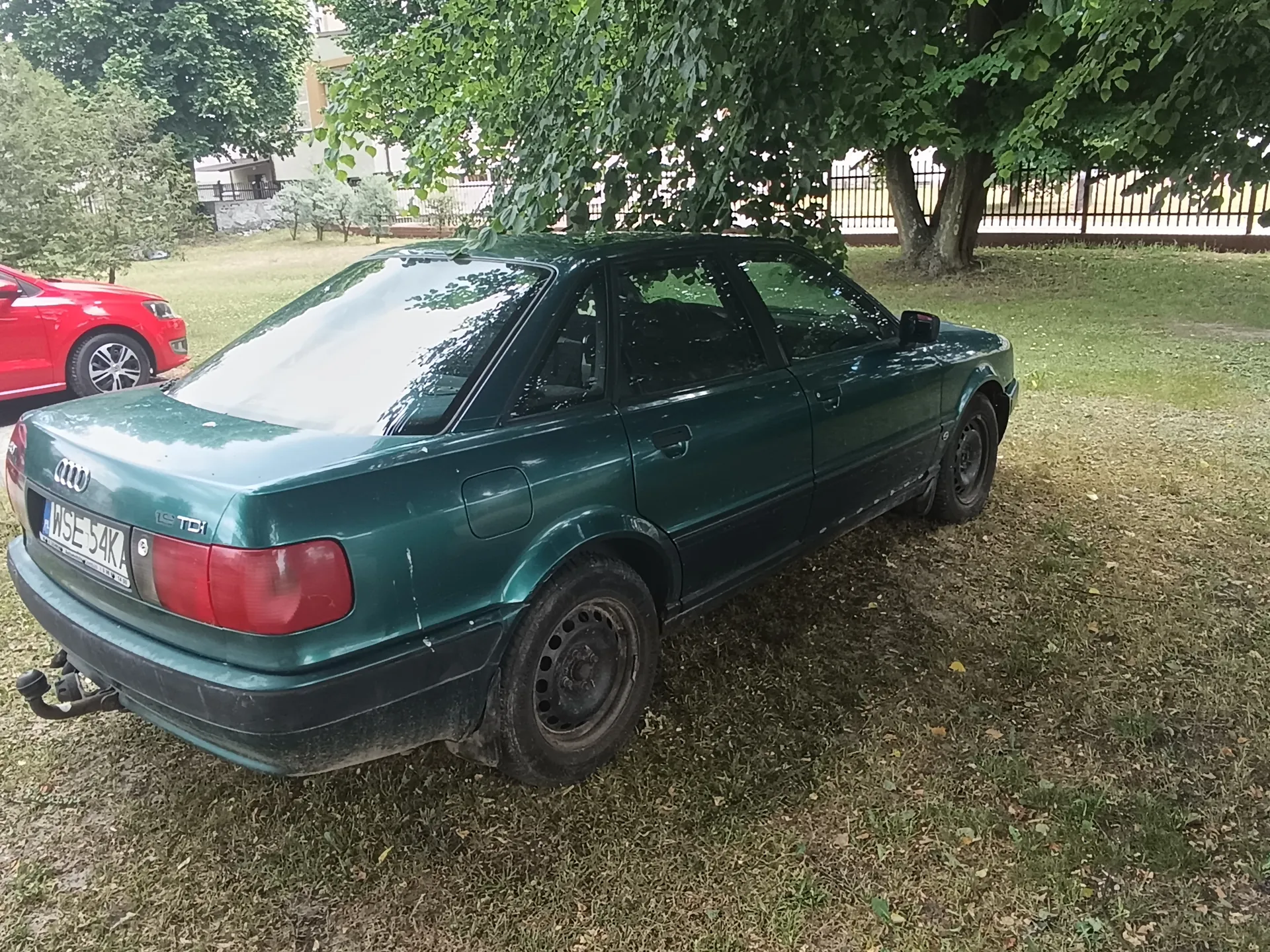 Zielony samochód marki Audi z tablicą rejestracyjną WSE 54KA i napisem 1.9 TDI na bagażniku, zaparkowany na trawie. W tle czerwony samochód, drzewa, metalowe ogrodzenie oraz budynek. Samochód ma czarne felgi i widoczne ślady zużycia na karoserii.