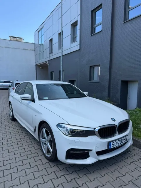 Biały samochód marki BMW zaparkowany na kostce brukowej przed nowoczesnym budynkiem o szarej i białej elewacji. Samochód ma polską tablicę rejestracyjną o numerze SO 7006W.