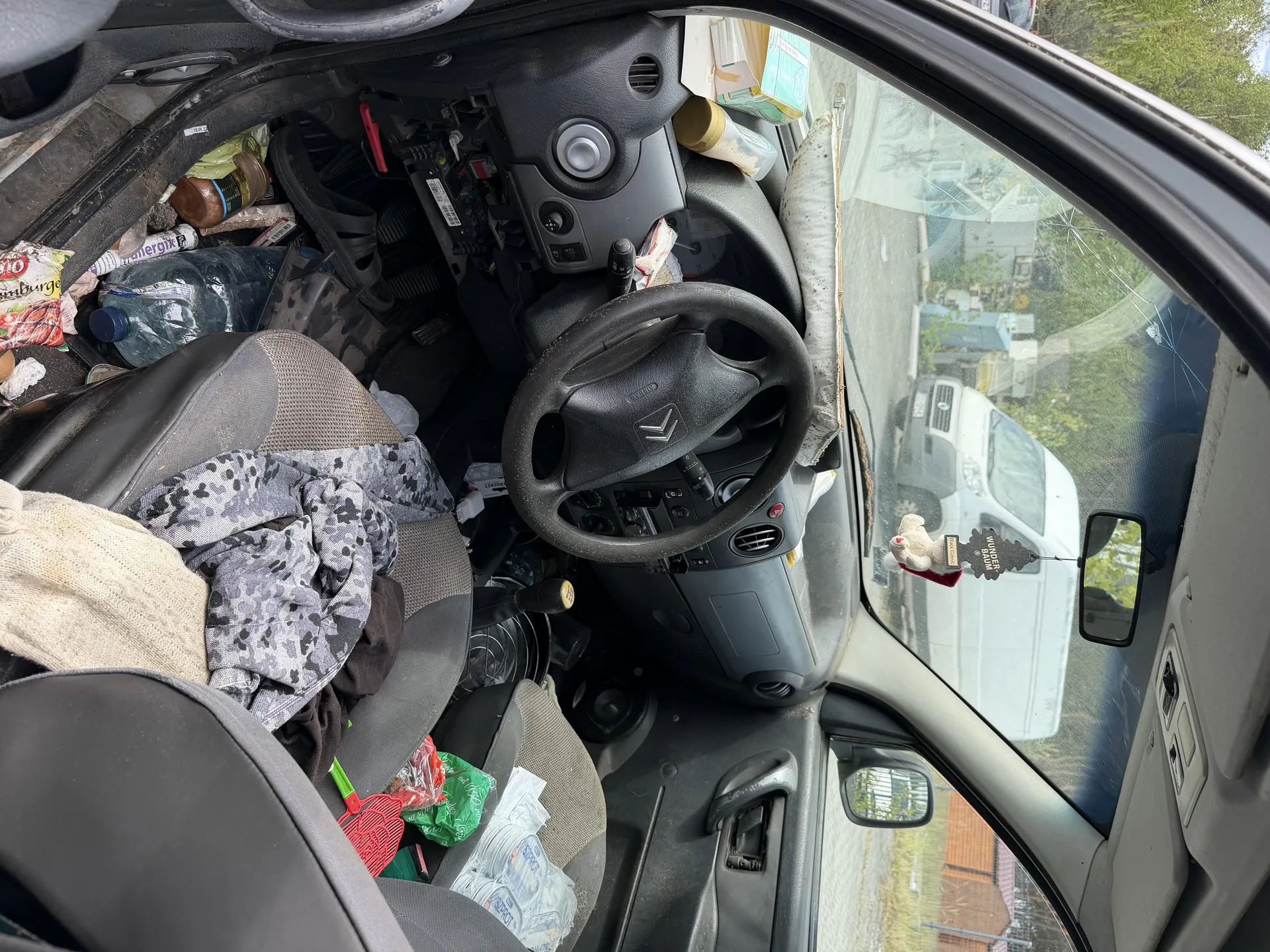 Wnętrze samochodu w stanie silnego zanieczyszczenia. Widoczna kierownica z logo Citroëna, rozrzucone przedmioty na siedzeniach: ubrania (w tym szara tkanina w kamuflażu i beżowy sweter), plastikowe butelki, opakowania po jedzeniu, czerwony przedmiot przypominający szczotkę. Na desce rozdzielczej leżą różne przedmioty, w tym butelka i opakowanie. Przez przednią szybę widać biały samochód dostawczy i budynki. Na lusterku wstecznym wiszą ozdoby: pluszowy niedźwiedź i czarna tabliczka z napisem. Szyba przednia ma pęknięcie.