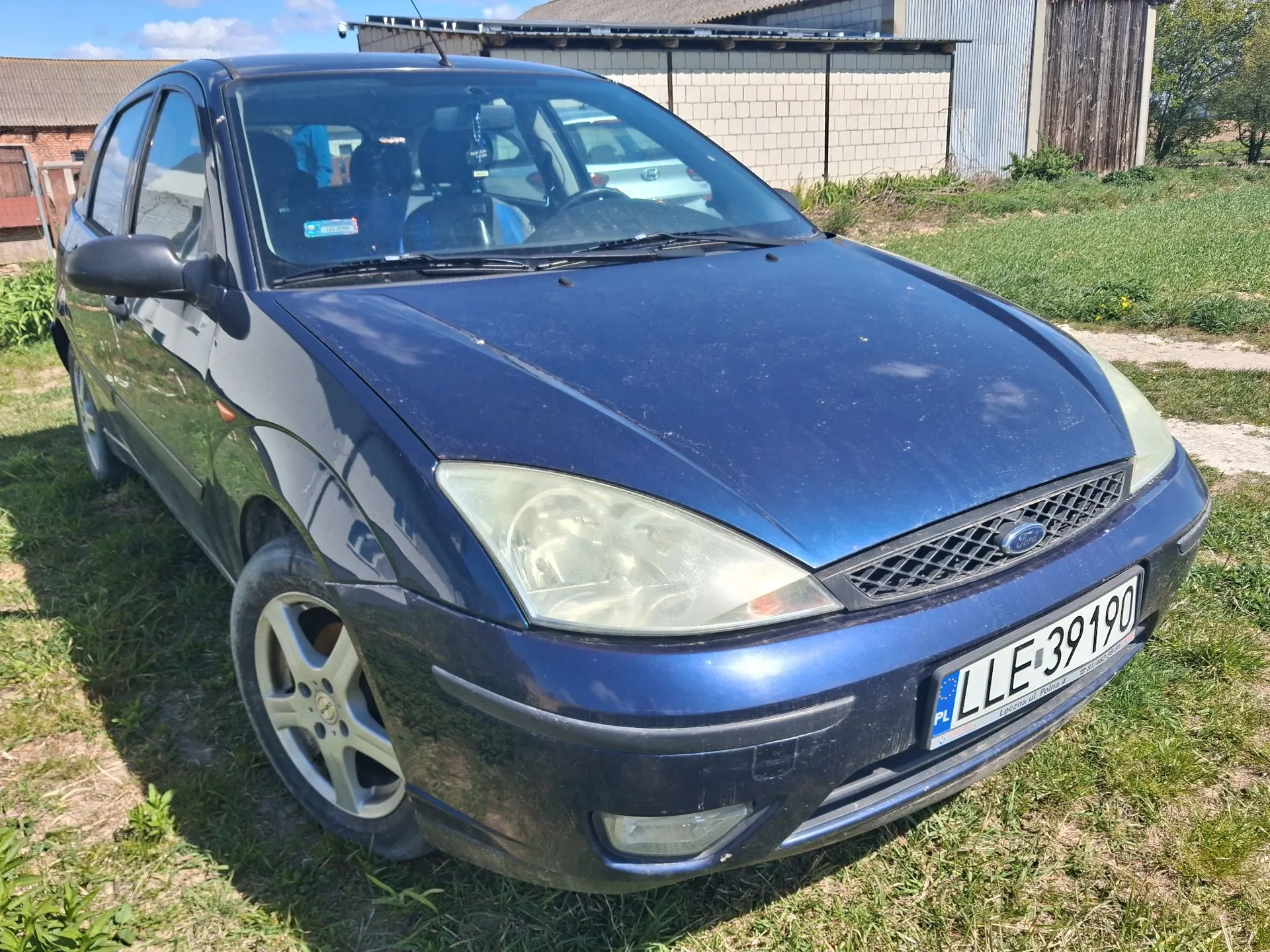Granatowy samochód osobowy marki Ford zaparkowany na trawiastym terenie. Pojazd jest widoczny z przodu pod kątem, z widoczną lewą stroną. Samochód ma zmatowione reflektory i polską tablicę rejestracyjną o numerze LLE 39190. W tle widoczny jest budynek z jasnej cegły oraz drewniany szop.