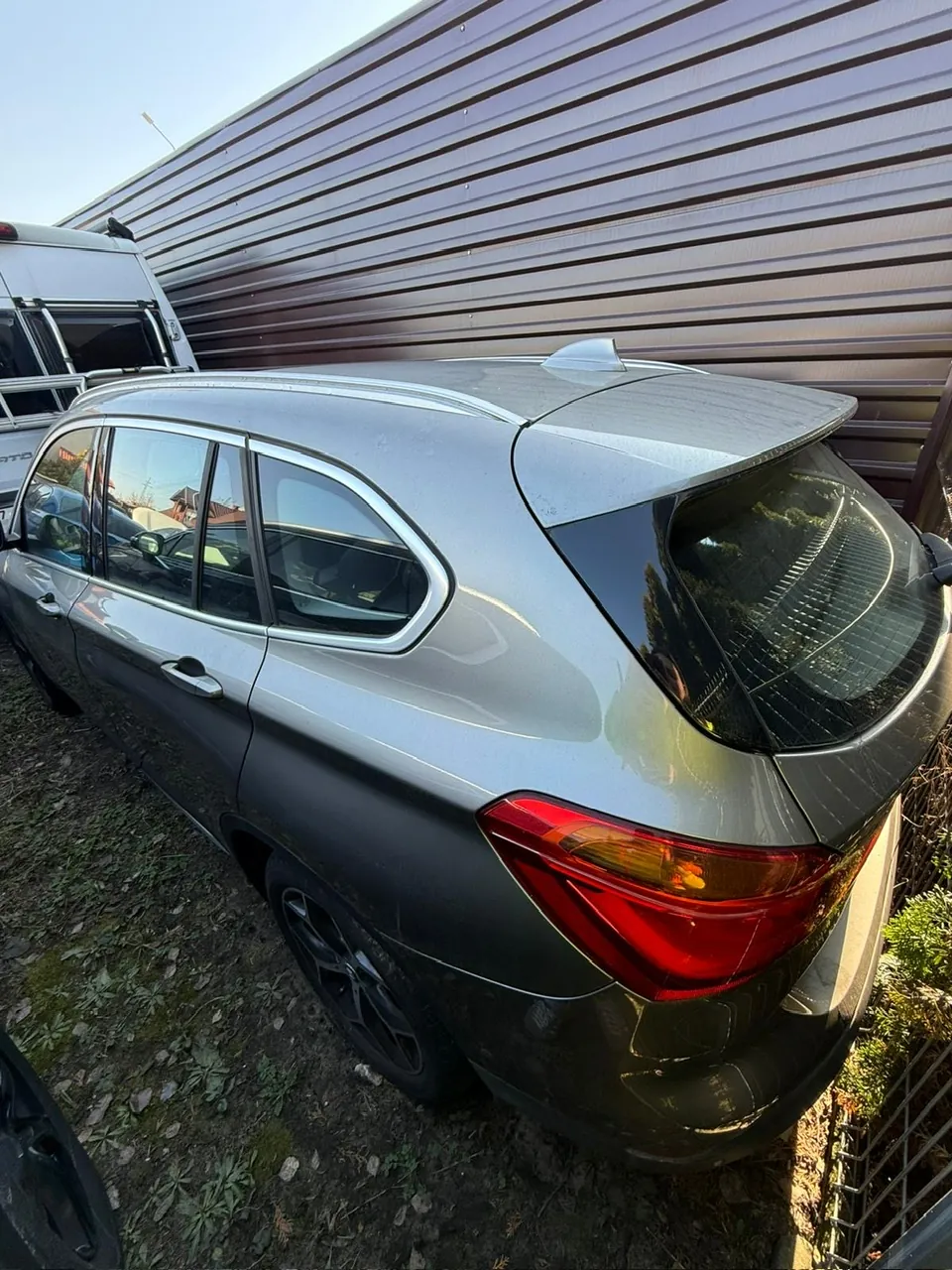 Srebrny samochód typu SUV zaparkowany na terenie z ziemią i trawą obok budynku z poziomymi metalowymi blachami. Widoczna tylna część pojazdu z czerwonymi światłami tylnymi i szybą. Po lewej stronie częściowo widoczny biały pojazd. Tło z jasnym niebem.