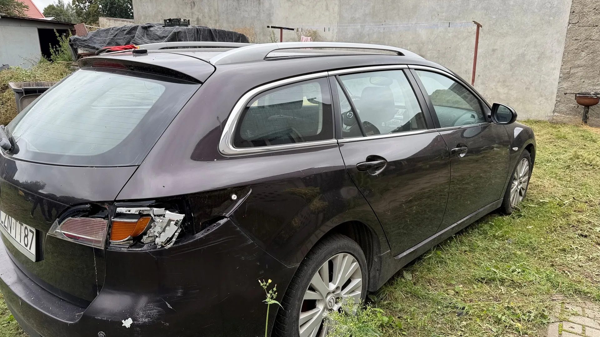Tylny i boczny widok ciemnofioletowego samochodu typu kombi zaparkowanego na trawie. Pojazd posiada widoczne uszkodzenia karoserii w tylnej części po lewej stronie, w tym rozbitą lampę tylną. Widoczny fragment tablicy rejestracyjnej z literami ZN i liczbami 187.