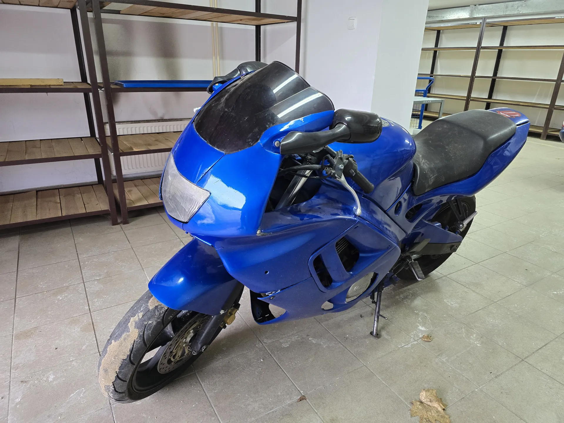Niebieski motocykl sportowy zaparkowany w pomieszczeniu z płytkami na podłodze. Motocykl ma czarną siedziskę, przednią szybę i zanieczyszczony przedni oponę. W tle widoczne są metalowe półki z drewnianymi blatami oraz biała ściana. Na podłodze leżą suche liście.