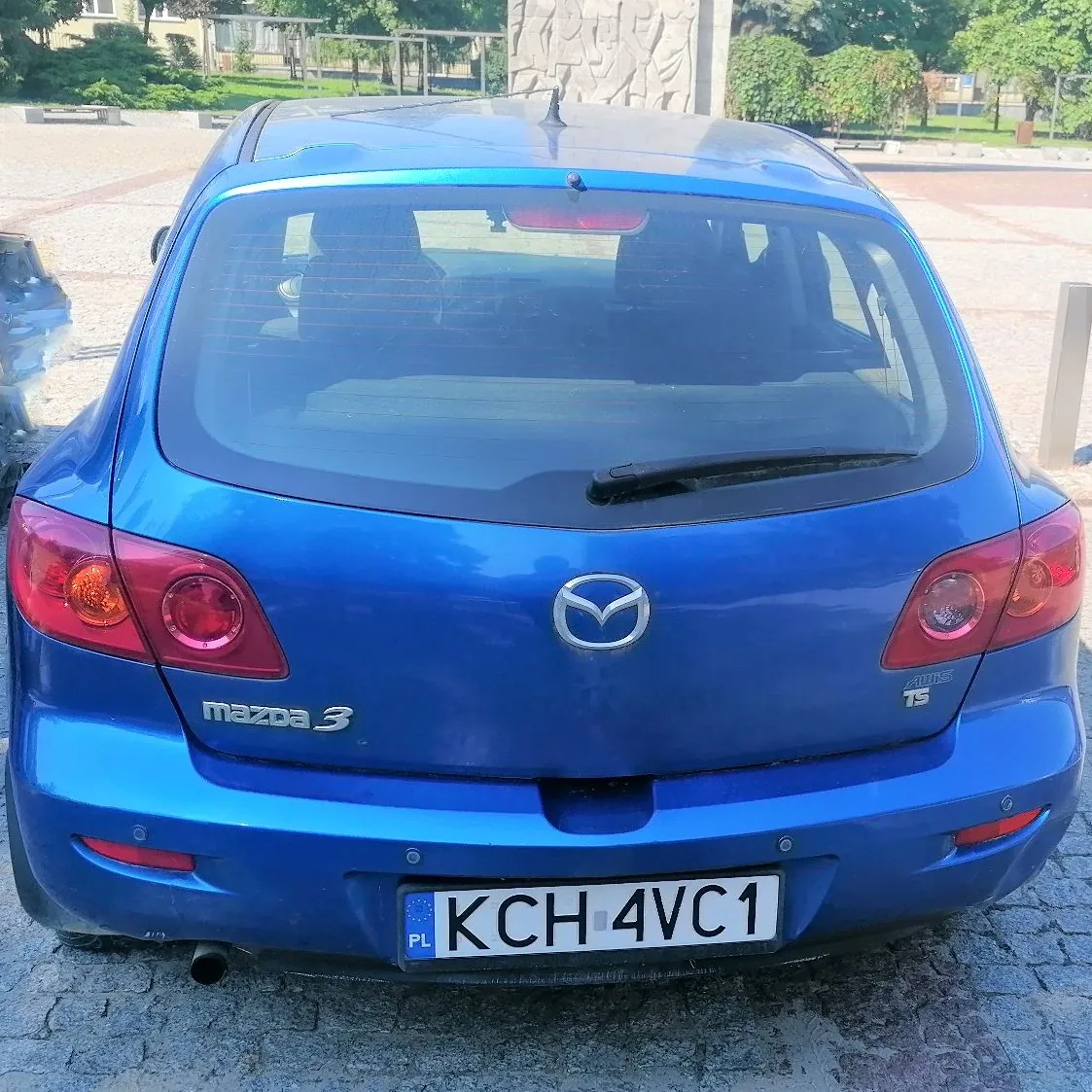 Tylny widok niebieskiego samochodu marki Mazda 3. Na klapie bagażnika znajduje się logo Mazdy oraz napis 'mazda 3' po lewej stronie i oznaczenie modelu '1.6 TS' po prawej. Samochód posiada polską tablicę rejestracyjną o numerze KCH 4VC1.