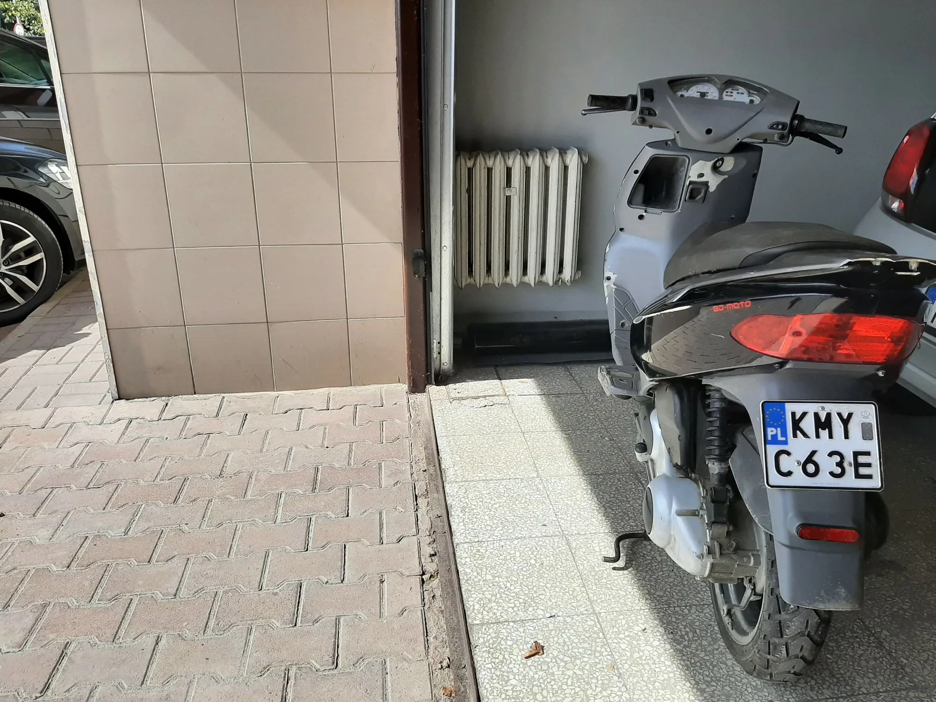 Zdjęcie przedstawiające tył szarego skutera zaparkowanego w garażu na płytkach. Skuter ma czarną kanapę i czerwoną lampę tylną. Widoczna jest polska tablica rejestracyjna o numerze KMY C63E. W tle widać biały grzejnik przy ścianie oraz fragment ciemnego samochodu po lewej stronie. Przed garażem znajduje się kostka brukowa.