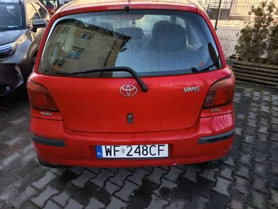 Czerwony samochód Toyota Yaris widziany od tyłu. Na tylnej szybie widoczne odbicie budynku. Na bagażniku umieszczono logo Toyoty oraz napis YARIS po prawej stronie. Tablica rejestracyjna z polskim kodem PL i numerem WF-248CF. Samochód zaparkowany na brukowanej powierzchni. Po lewej stronie częściowo widoczny inny pojazd, w tle ogrodzenie i krzewy.