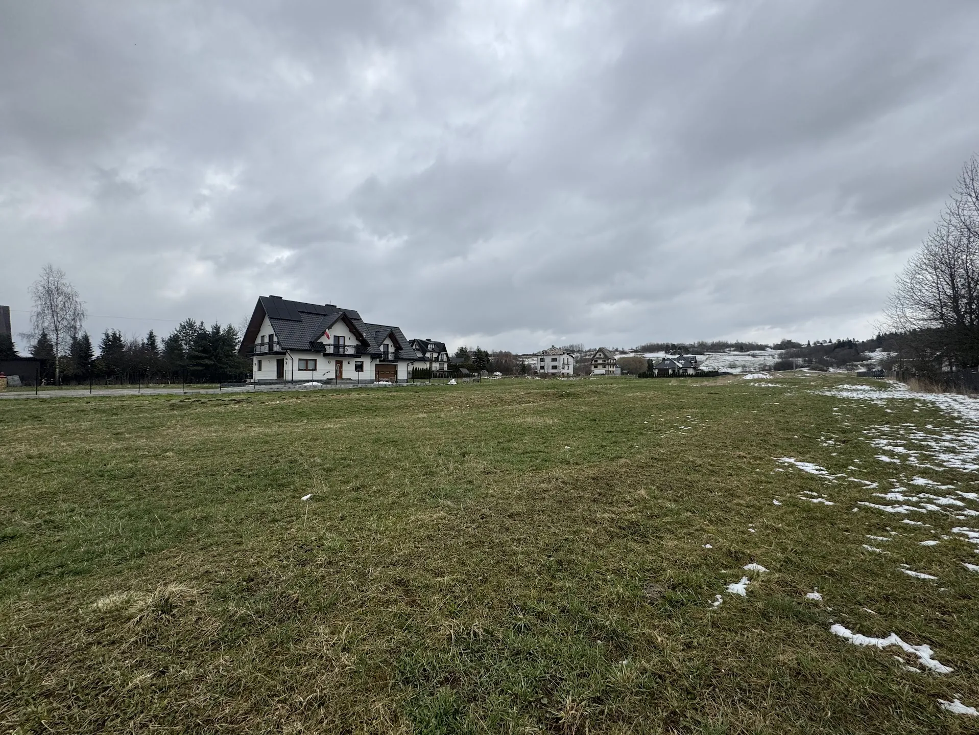 Rozległe pole trawiaste z resztkami śniegu po prawej stronie. W tle widoczne są domy o białych elewacjach i ciemnych dachach, otoczone drzewami. Niebo jest zachmurzone, z gęstymi szarymi chmurami. W oddali znajdują się kolejne budynki i lekko zalesiony teren.