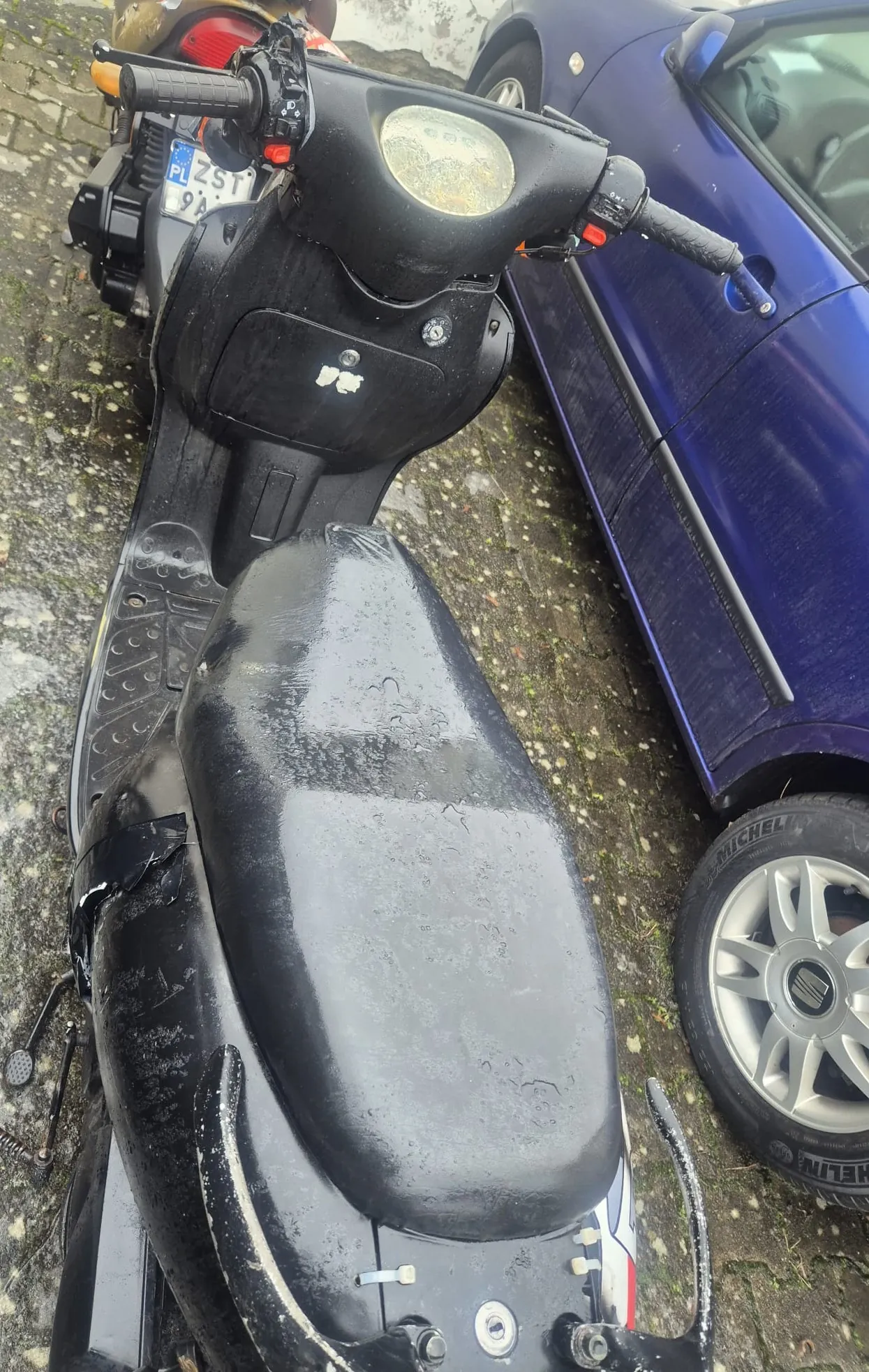 Czarny skuter z okrągłym reflektorem i czarną siedziskiem pokrytym kroplami wody, zaparkowany na brukowanej powierzchni. Obok niego po prawej stronie niebieski samochód z widocznym kołem z srebrną felgą i oponą Michelin. W tle drugi skuter z tablicą rejestracyjną PL ZST 9A. Na skuterze widoczne są ślady zużycia i mały biały znak na przedniej części.