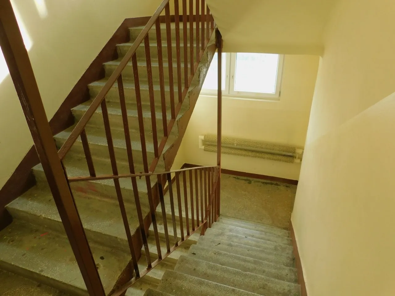 Widok na betonowe schody wewnątrz budynku z brązową, metalową balustradą. Schody prowadzą w górę i w dół. W tle widoczna jest jasna ściana z oknem oraz zamontowany pod nim poziomy grzejnik.