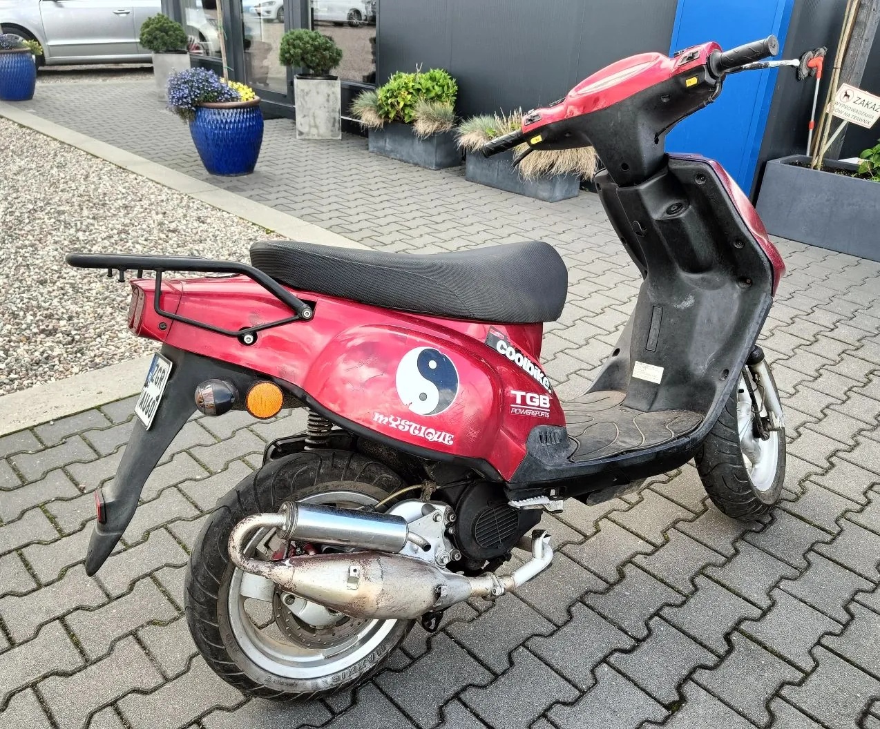 Czerwony skuter marki TGB z czarnym siedzeniem, zaparkowany na kostce brukowej. Na boku skutera znajduje się biały symbol yin-yang oraz napisy 'TGB POWERSPORTS' i 'mystique'. Pojazd posiada metalowy bagażnik z tyłu i srebrny wydech. W tle widoczny jest szary budynek, niebieskie donice z kwiatami oraz fragment srebrnego samochodu.