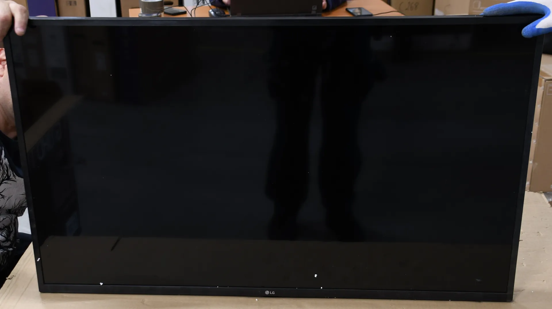 Czarny, wyłączony ekran telewizora marki LG z cienkimi ramkami, trzymany przez dwie osoby. Na ekranie widoczne jest odbicie sylwetki osoby stojącej naprzeciwko.