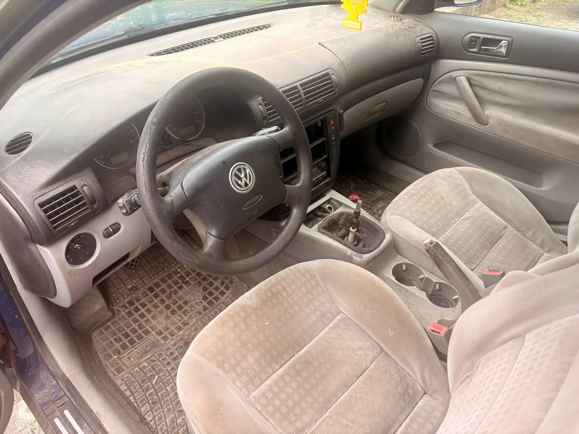 Wnętrze samochodu marki Volkswagen z perspektywy pasażera. Widoczna jest zakurzona deska rozdzielcza, kierownica z logo VW, dwa przednie fotele z jasnej tkaniny oraz gumowe dywaniki na podłodze. W centralnej konsoli znajduje się dźwignia zmiany biegów i uchwyty na kubki.