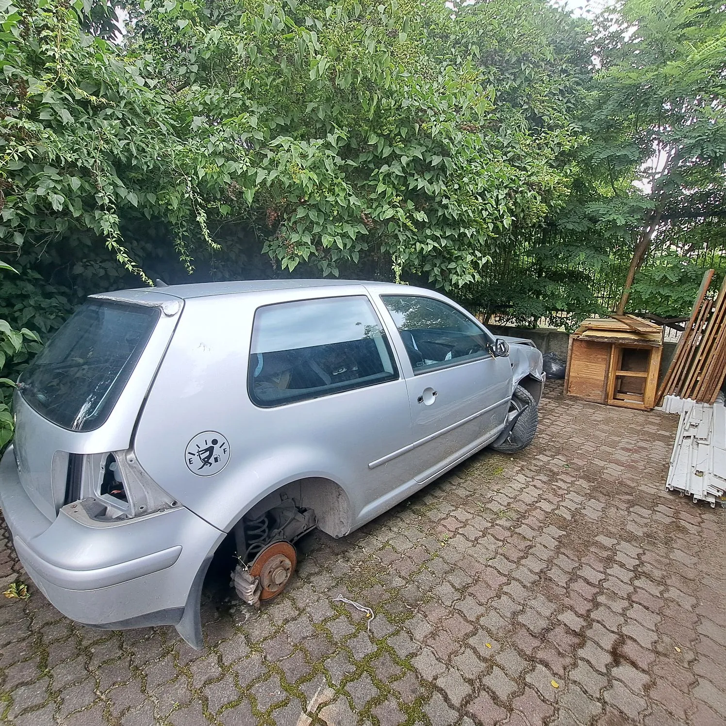 Srebrny samochód osobowy typu hatchback, pozbawiony kół, stojący na kostce brukowej. Pojazd jest uszkodzony, brakuje mu tylnego lewego reflektaru oraz obu kół z lewej strony, odsłaniając elementy zawieszenia i tarcze hamulcowe. Na tylnym błotniku znajduje się okrągła naklejka z grafiką. W tle widoczne są gęste, zielone krzewy i drzewa oraz drewniana konstrukcja przypominająca budę dla psa.
