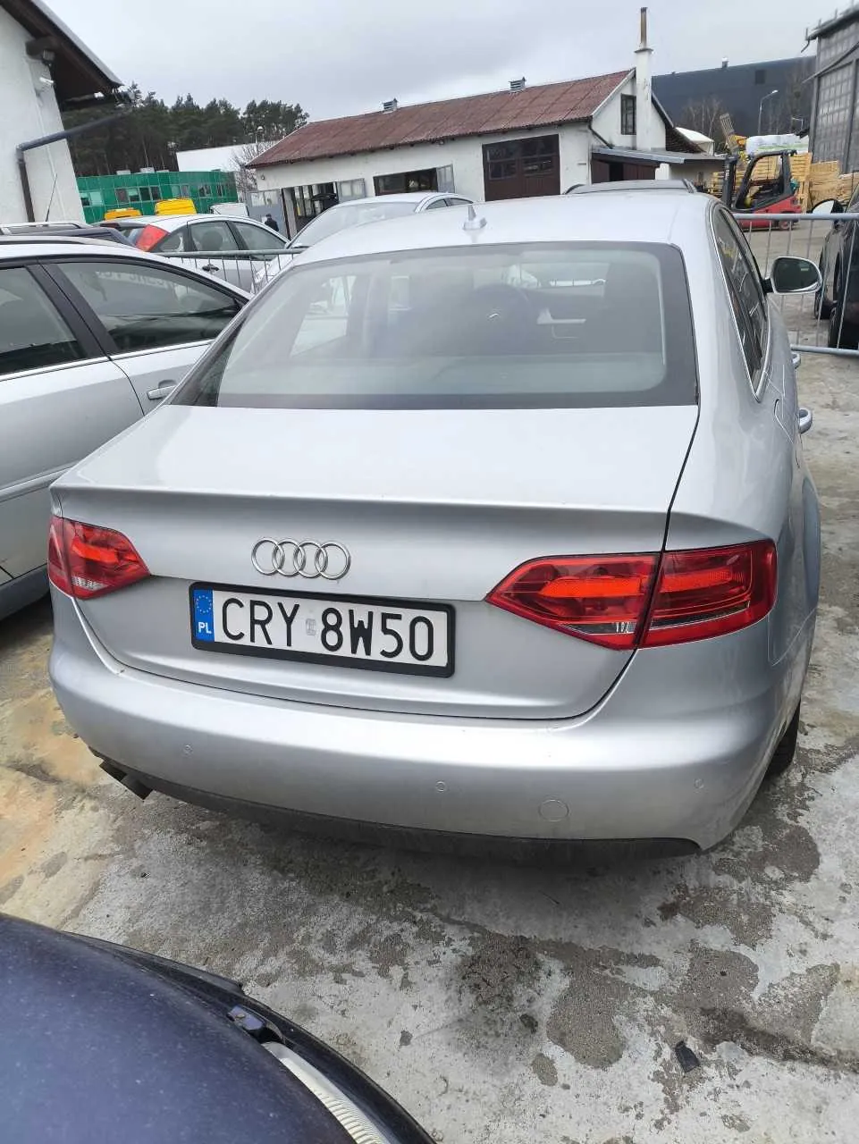 Srebrny samochód Audi z tyłu, z widoczną tablicą rejestracyjną CRY 8W50 z flagą PL. W tle inne pojazdy, białe budynki z brązowym dachem oraz betonowa powierzchnia z plamami.