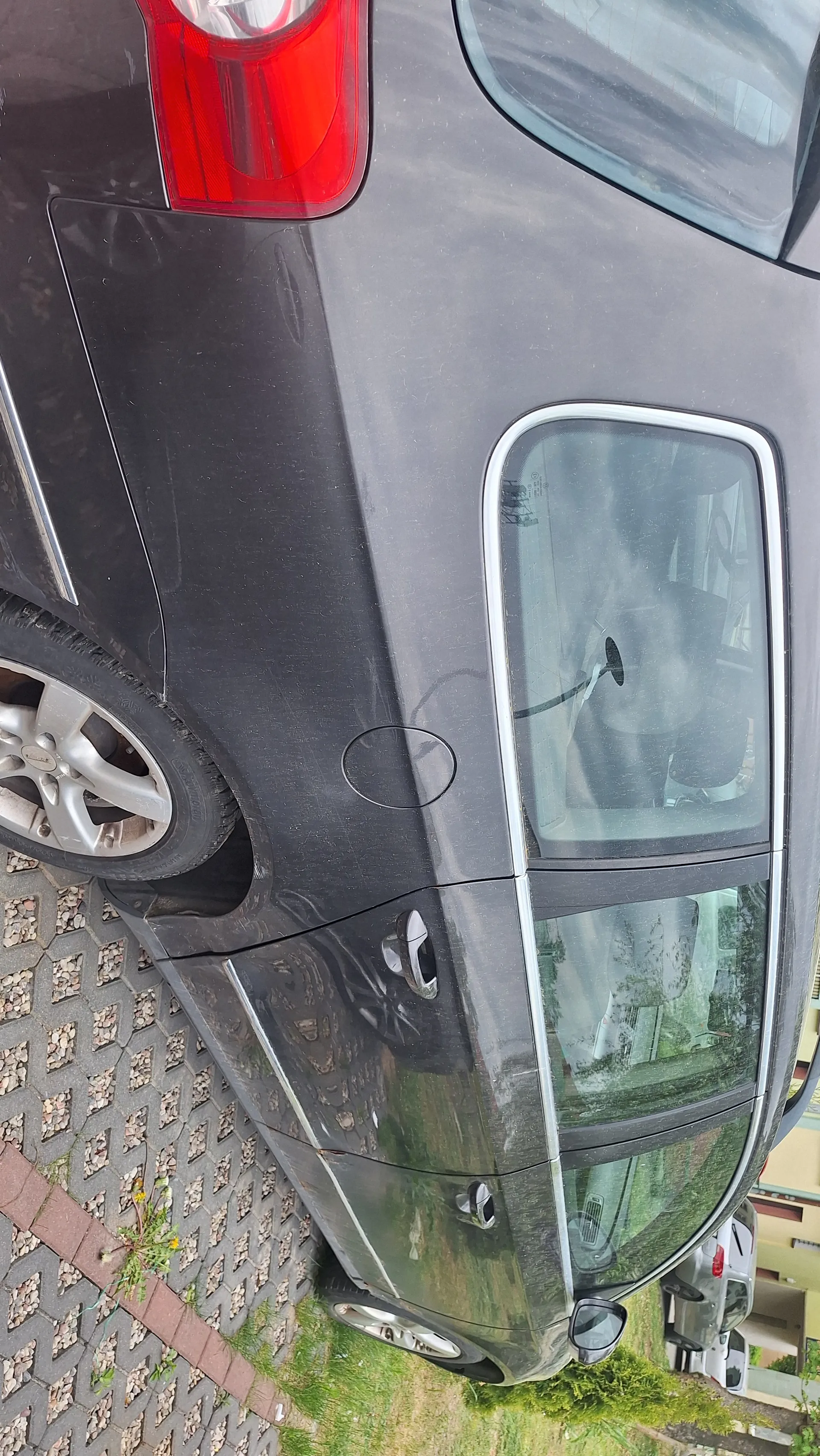 Ciemnoszary samochód widziany z góry, zaparkowany na brukowanej powierzchni. Widoczne są tylny reflektor, koło z felgą, boczne okna oraz fragment przedniej części. W tle trawa i fragment innych pojazdów.