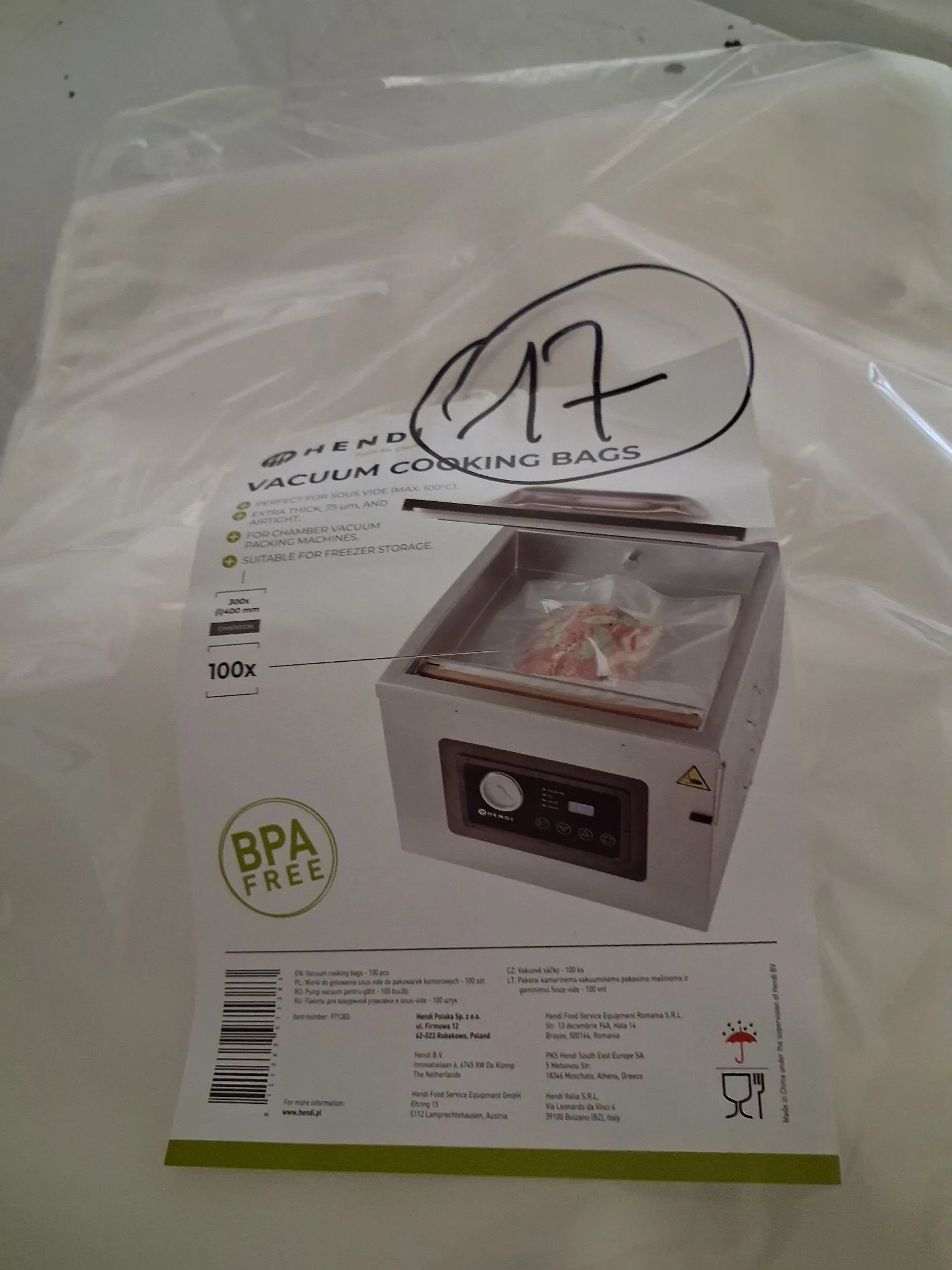 Zdjęcie opakowania folii do gotowania próżniowego marki Hend. Na białym opakowaniu widnieje napis 'VACUUM COOKING BAGS' oraz lista cech produktu: 'PERFECT FOR SOUS VIDE (MAX. 100°C)', 'EXTRA THICK, 75 µm, AND AIRTIGHT', 'FOR CHAMBER VACUUM PACKING MACHINES' oraz 'SUITABLE FOR FREEZER STORAGE'. Widoczna jest grafika przedstawiająca pakowarkę próżniową z włożonym do niej woreczkiem z mięsem. Na opakowaniu znajduje się logo 'BPA FREE', informacja o ilości '100x' oraz wymiarach '300x400 mm'. W prawym górnym rogu widnieje ręcznie napisana liczba '17' obwiedziona kółkiem. Na dole znajdują się dane adresowe dystrybutorów z różnych krajów, w tym z Polski (Hend Polska Sp. z o.o.).