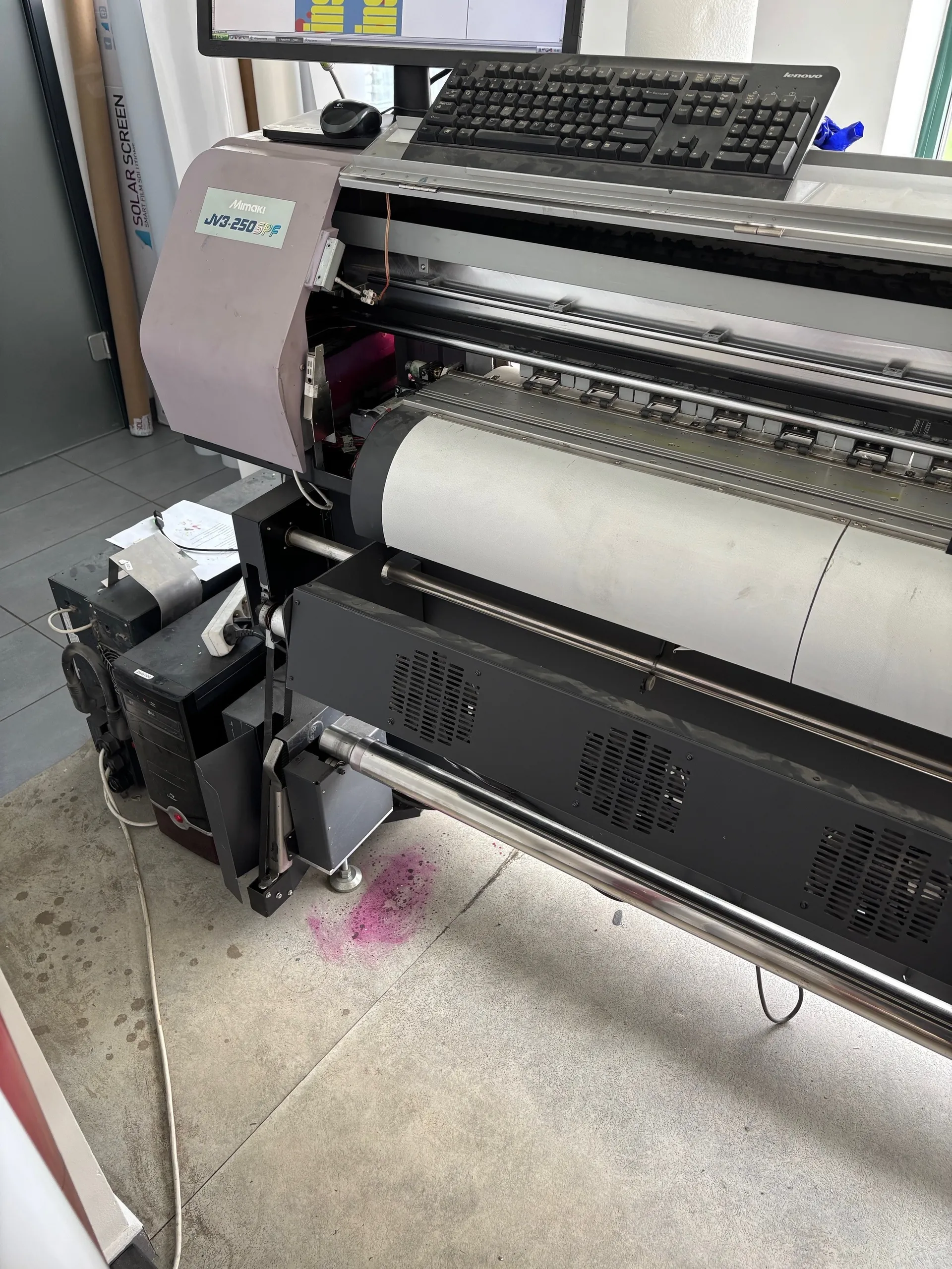 Duży drukarka ploterowa marki Mimaki model JV3-250SPF z rolką białego materiału. Na drukarce umieszczono czarną klawiaturę Lenovo, mysz oraz monitor. Obok drukarki znajduje się czarny komputer stacjonarny z przewodami. Na podłodze widoczna różowa plama. W tle rulon z napisem 'SOLAR SCREEN' oraz fragment okna.