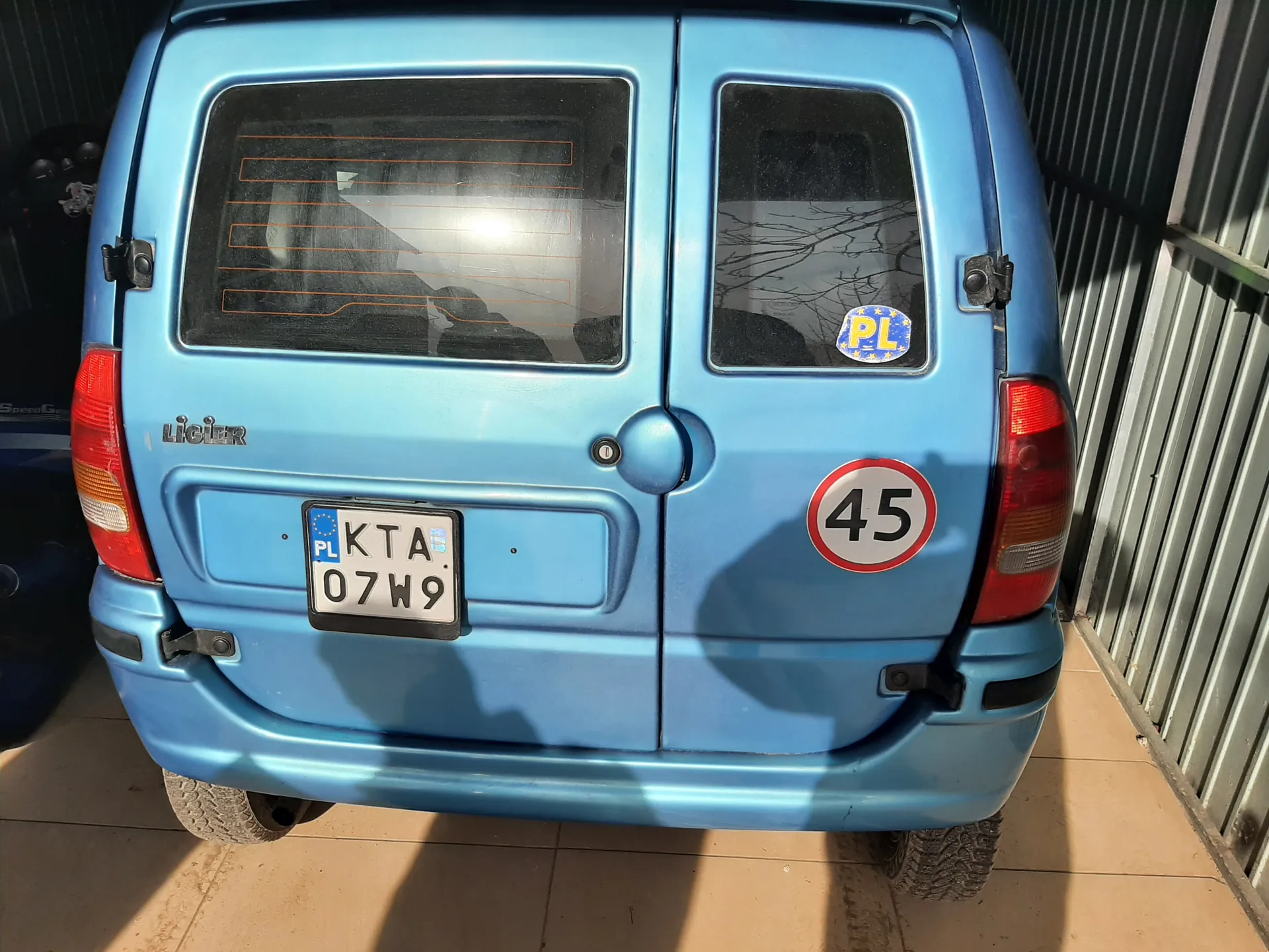 Tył niebieskiego samochodu dostawczego marki Ligier. Widoczna tablica rejestracyjna PL KTA 07W9, okrągły znak z cyfrą 45 oraz naklejka PL z gwiazdkami. Samochód stoi na podłodze z płytek, obok metalowej ściany. W tle odbicie gałęzi drzew w szybie.