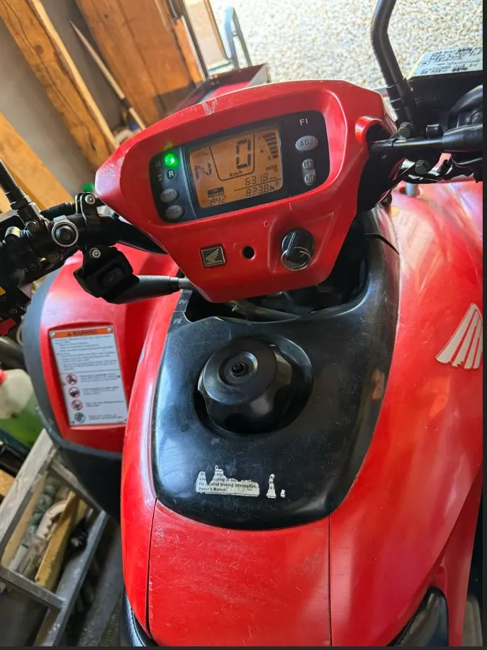Zbliżenie na kokpit czerwonego quada marki Honda. Widoczny jest cyfrowy wyświetlacz z prędkościomierzem wskazującym 0 km/h, licznikiem przebiegu 823.8 hm oraz wskaźnikiem paliwa. Na panelu znajduje się logo Hondy oraz przyciski sterujące. Poniżej wyświetlacza widoczny jest czarny korek wlewu paliwa osadzony w czerwonej obudowie pojazdu. Po lewej stronie znajduje się biała naklejka ostrzegawcza z piktogramami.
