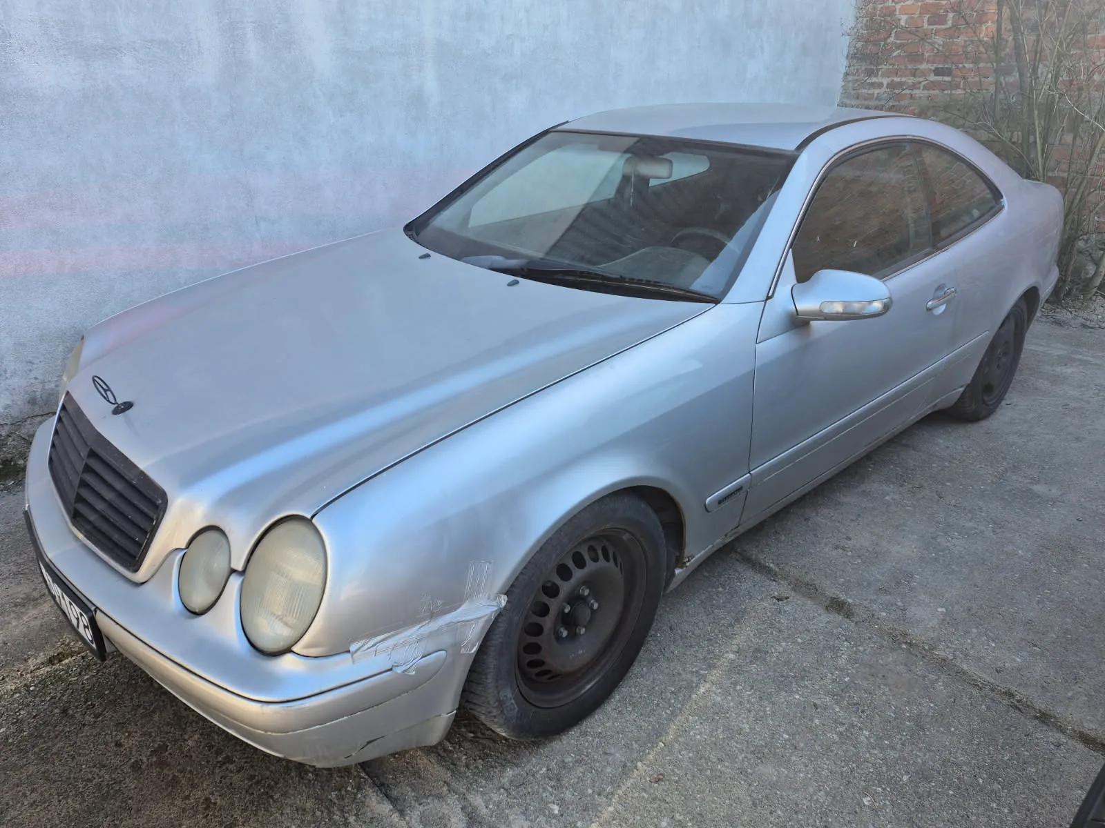 Srebrny samochód osobowy marki Mercedes-Benz typu coupe zaparkowany na betonowej powierzchni. Widoczne są uszkodzenia przedniego zderzaka z taśmą naprawczą, zużyte felgi oraz częściowo widoczna tablica rejestracyjna. W tle betonowa i ceglana ściana oraz suche krzewy. Samochód posiada charakterystyczne okrągłe reflektory i logo marki na masce.