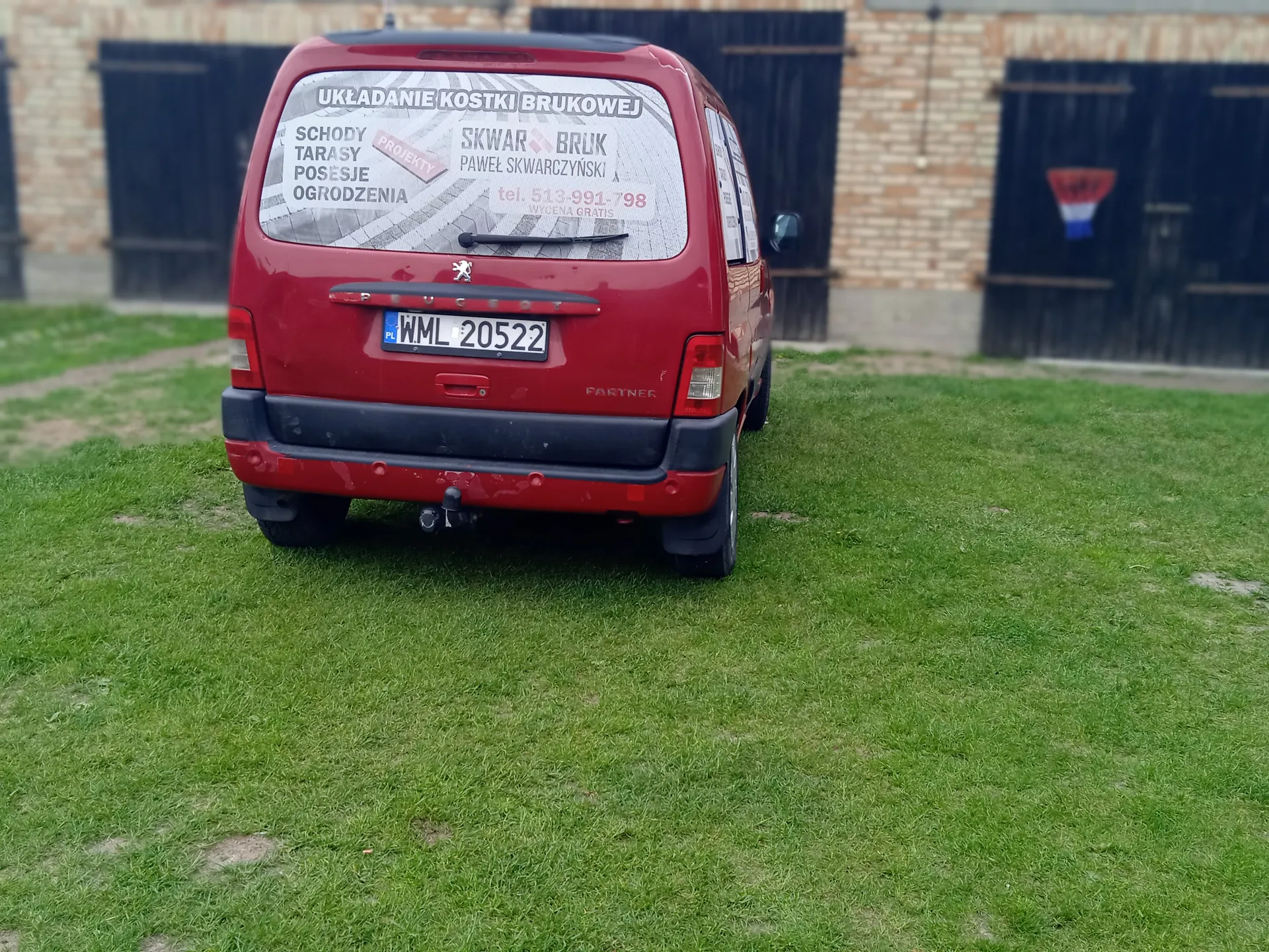 Czerwony samochód dostawczy Peugeot Partner zaparkowany tyłem na trawie. Na tylnej szybie widoczna reklama z napisami: "UKŁADANIE KOSTKI BRUKOWEJ", "SCHODY TARASY POSESJE OGRODZENIA", "SKWAR BRUK PAWEŁ SKWARCZYŃSKI", "tel. 513-991-798" oraz tablica rejestracyjna "WML 20522". W tle ceglany budynek z czarnymi drzwiami garażowymi i trawą na pierwszym planie.