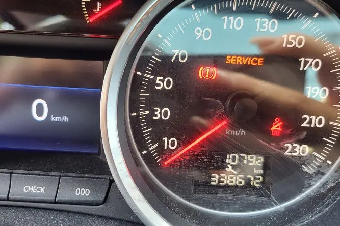 Zbliżenie na deskę rozdzielczą samochodu. Po prawej stronie znajduje się okrągły prędkościomierz z cyframi od 10 do 230 km/h. Wskazówka prędkościomierza znajduje się na poziomie 0. Na tarczy widnieje pomarańczowy napis „SERVICE” oraz czerwona kontrolka układu hamulcowego. W dolnej części prędkościomierza znajduje się cyfrowy wyświetlacz z liczbami 1079.2 km oraz 33867.2. Po lewej stronie widoczny jest fragment ekranu z cyfrą „0 km/h” oraz przyciski „CHECK” i „000”.
