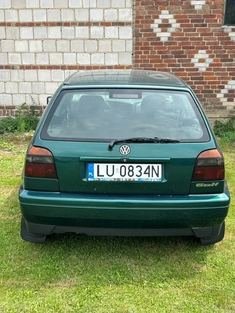 Tylny widok ciemnozielonego samochodu Volkswagen Golf zaparkowanego na trawie przed ścianą z białych i czerwonych cegieł. Samochód ma polską tablicę rejestracyjną o numerze LU 0834N oraz logo Volkswagena na klapie bagażnika.