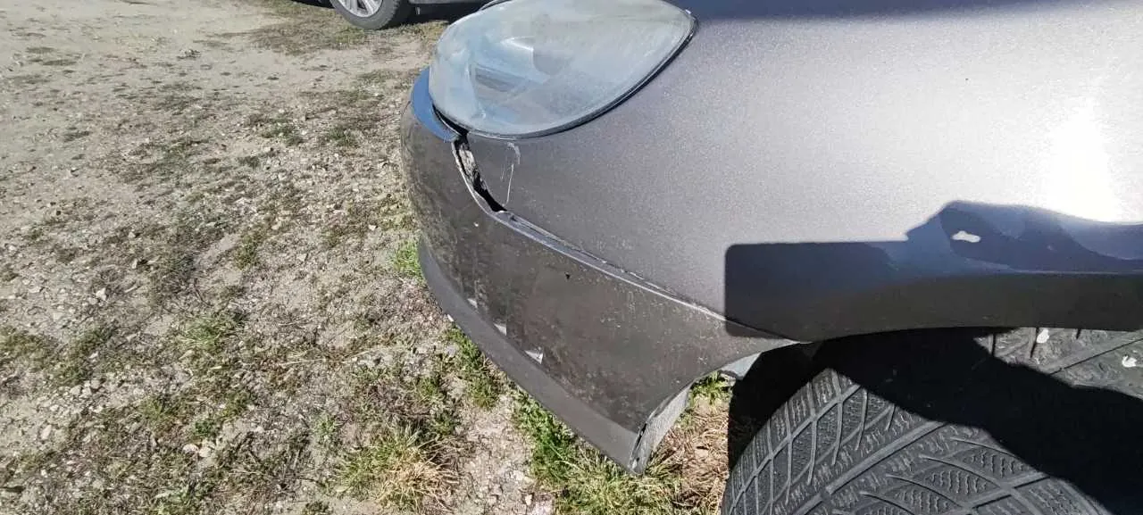 Zbliżenie na przód szarego samochodu z uszkodzonym zderzakiem i reflektorem. Widoczne pęknięcie w zderzaku blisko reflektora. Tło stanowi ziemia z rzadką trawą i kamieniami. W tle częściowo widoczne koło innego pojazdu. Brak czytelnego tekstu.