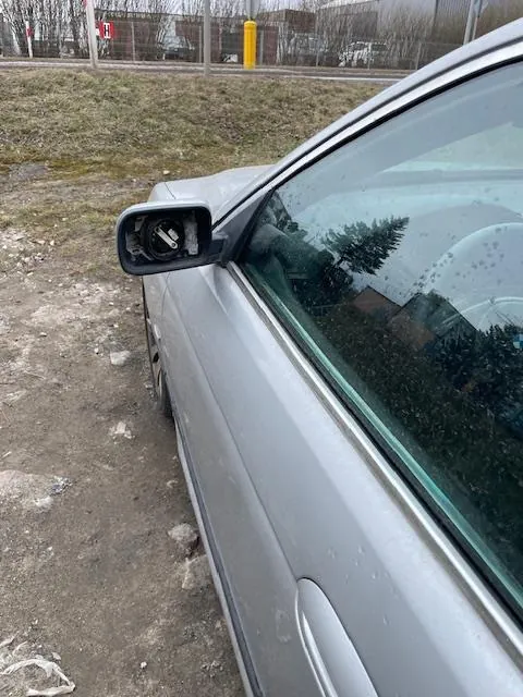 Srebrny samochód z uszkodzonym lusterkiem bocznym, zaparkowany na terenie z brudną ziemią i trawą. W tle widoczne są trawiaste obszary, żółty słup, drzewa oraz budynki.