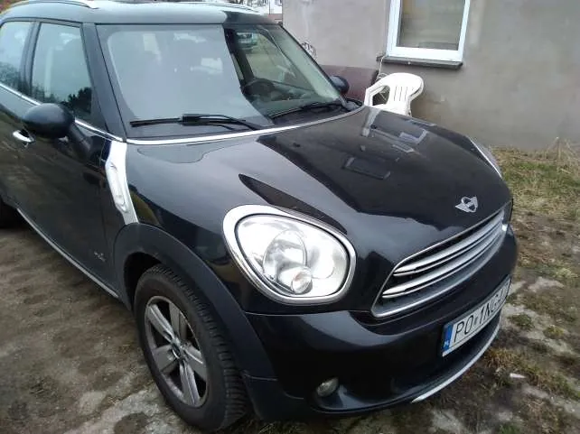 Czarny samochód Mini Cooper zaparkowany na podwórku. Widoczna tablica rejestracyjna z napisem PO 1NG. W tle budynek z oknem oraz białe krzesło plastikowe.