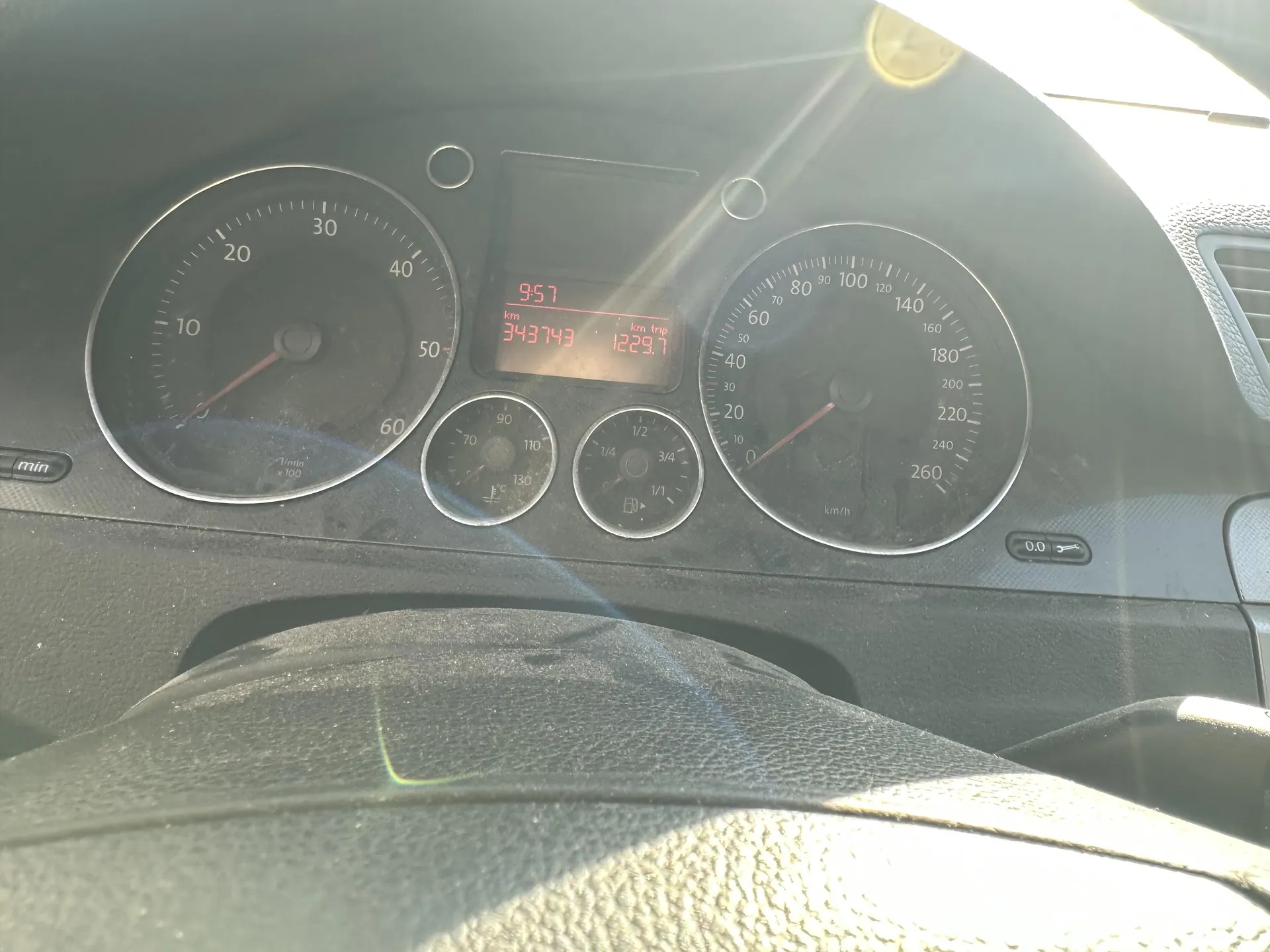 Zbliżenie na deski rozdzielcze samochodu. Po lewej stronie wskaźnik obrotomieru z zakresem 0-60 (x1000 obr./min), po prawej prędkościomierz z zakresem 0-260 km/h. W centrum wyświetlacz cyfrowy z czerwonymi cyframi: godzina 9:57, przebieg 343743 km, przebieg podróży 1229.7 km. Poniżej wyświetlacz znajdują się dwa mniejsze wskaźniki: termometr (zakres do 130) i wskaźnik paliwa (podziałki 1/4, 1/2, 3/4, 1/1). Na desce widoczne są również napisy: 'min' po lewej stronie, 'km/h' pod prędkościomierzem oraz '0.0' po prawej stronie. W dolnej części kadru widoczna część kierownicy. Deska rozdzielcza ma czarny kolor z srebrnymi obramowaniami wskaźników. Na obrazie widoczne są refleksy światła słonecznego.
