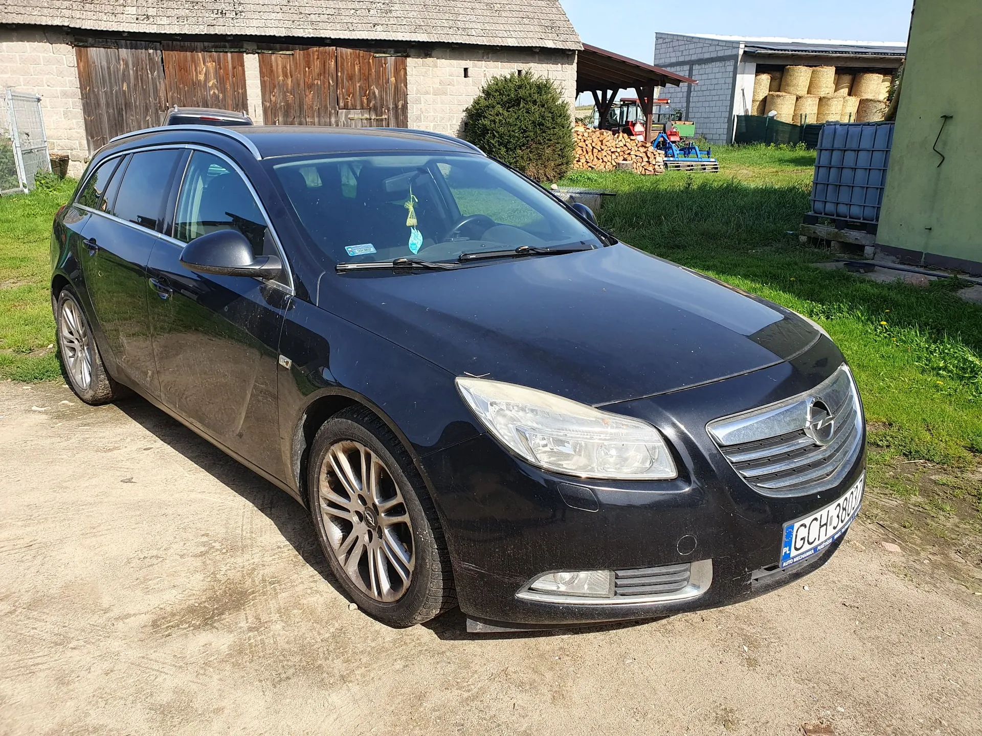 Czarny samochód osobowy typu kombi zaparkowany na piaszczystej drodze. W tle widoczny jest drewniany budynek gospodarczy, zielona trawa oraz stóg siana w oddali. Samochód ma tablicę rejestracyjną o numerze GCH 3803.