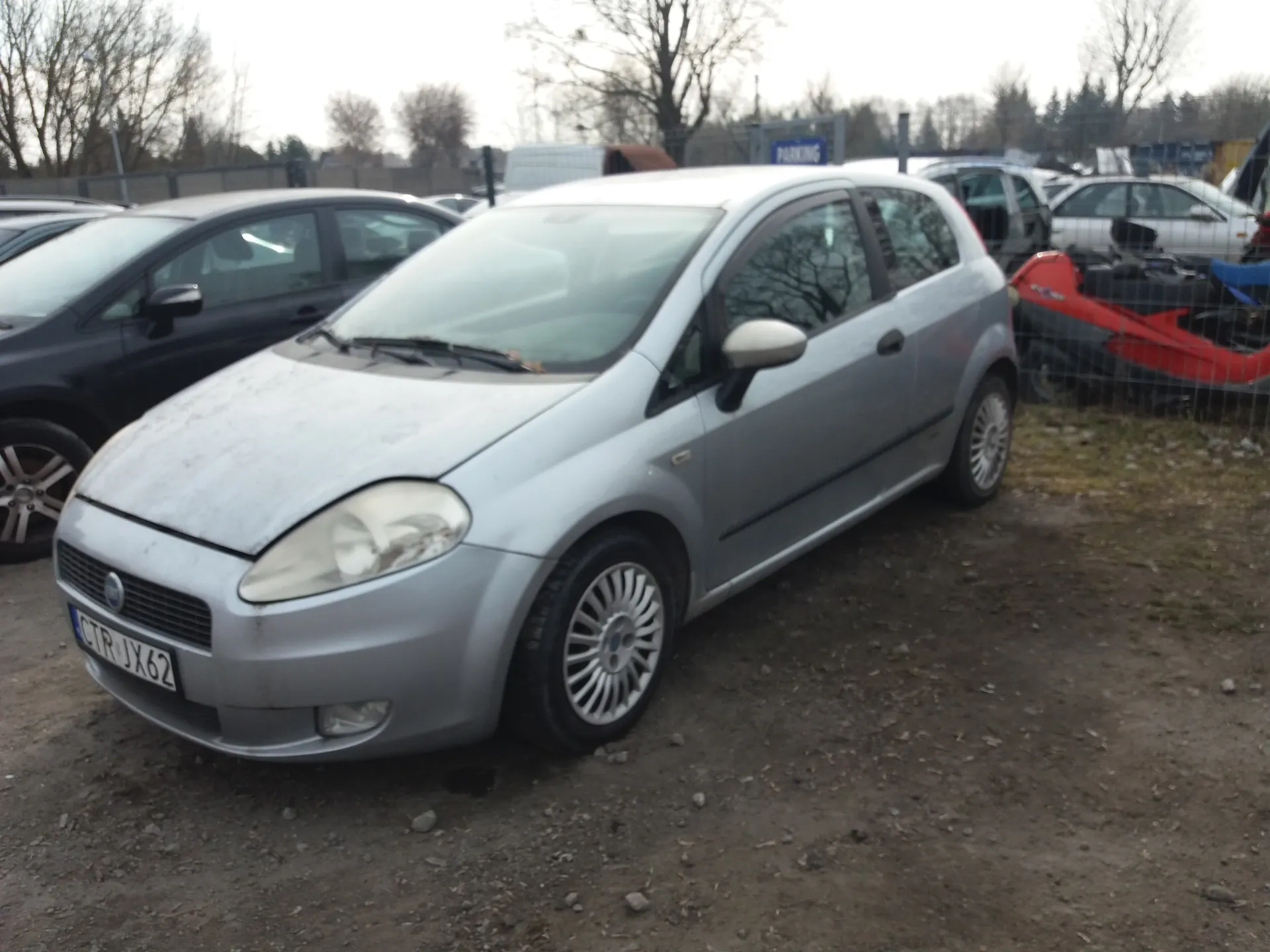 Srebrny samochód osobowy typu hatchback z tablicą rejestracyjną CTR JX62 zaparkowany na żwirowym terenie. W tle widoczne są inne pojazdy, w tym czarny samochód, biały samochód oraz czerwony skuter za ogrodzeniem. Na tle drzewa bez liści, niebieski znak z napisem PARKING oraz ogrodzenie. Teren otoczony jest drzewami i budynkami w oddali.