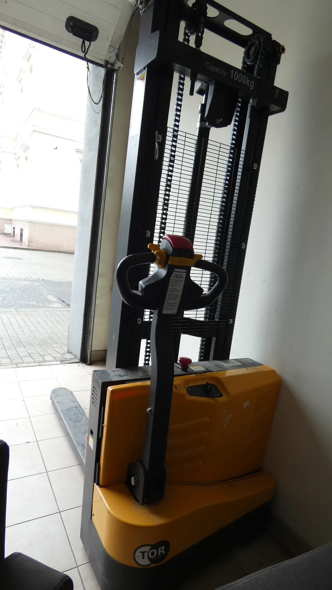 Żółto-czarny wózek podnośnikowy marki TOR z widocznym napisem 'Capacity: 1000kg' na czarnej ramie. Wózek posiada kierownicę z czerwonym i żółtym elementem oraz tabliczką z instrukcjami. W tle otwarte drzwi prowadzące na zewnątrz, gdzie widoczne są budynki i brukowana powierzchnia. Podłoga w pomieszczeniu wyłożona jest białymi płytkami, a po prawej stronie znajduje się biała ściana.