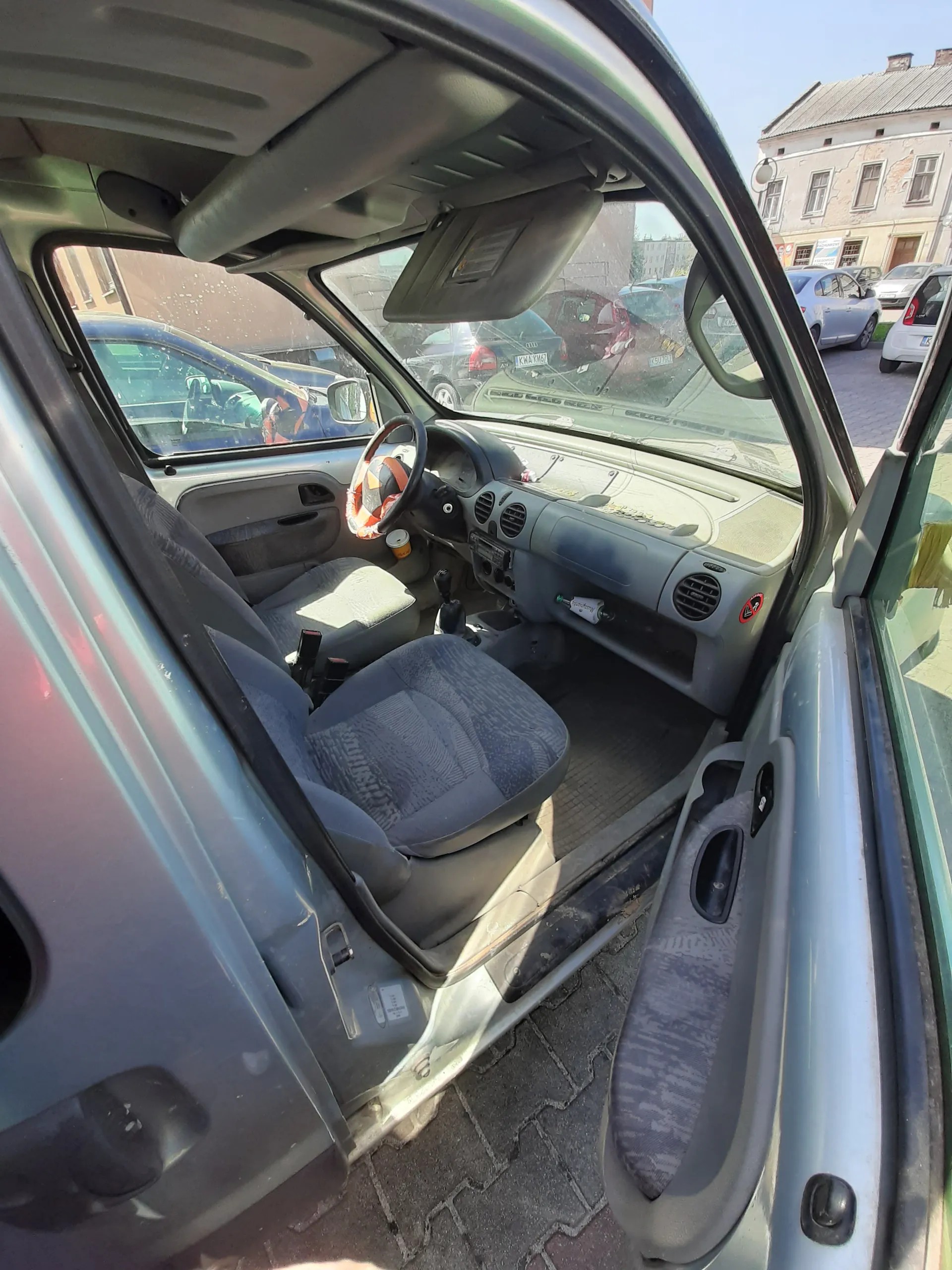 Wnętrze samochodu z otwartą drzwiami kierowcy. Widoczne są szare siedzenia z wzorem, kierownica z pomarańczową obiciem, deska rozdzielcza z wentylacjami i butelką. Poza samochodem widoczne są budynki i zaparkowane pojazdy.