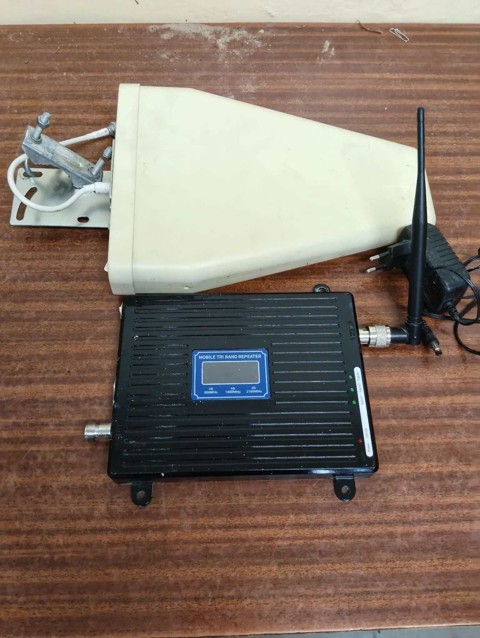 Zestaw urządzeń elektronicznych leżących na brązowym, drewnianym blacie. Na górze znajduje się biała, plastikowa obudowa o trapezoidalnym kształcie z zamontowanym metalowym uchwytem po lewej stronie. Poniżej znajduje się czarne, metalowe urządzenie z żebrowanym radiatorem na górnej obudowie. Na przednim panelu urządzenia widnieje napis 'MOBILE TRI BAND REPEATER', mały wyświetlacz LCD oraz oznaczenia pasm: '2G 900MHz', '3G 1800MHz' i '4G 2100MHz'. Do urządzenia podłączona jest czarna antena zewnętrzna oraz zasilacz sieciowy.