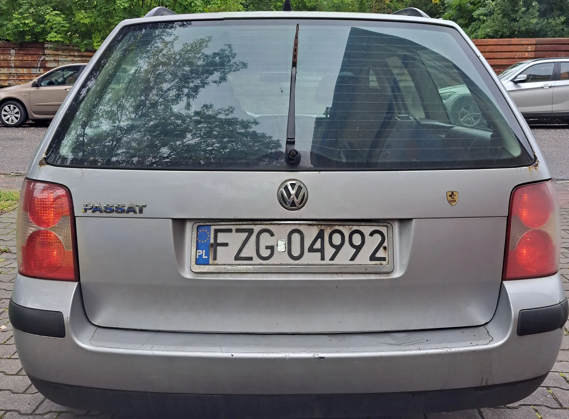 Tylny widok srebrnego samochodu marki Volkswagen Passat. Na klapie bagażnika widnieje napis 'PASSAT', logo Volkswagena oraz polska tablica rejestracyjna o numerze FZG 04992. Samochód ma tylną szybę z wycieraczką i dwa czerwone światła tylne po bokach.