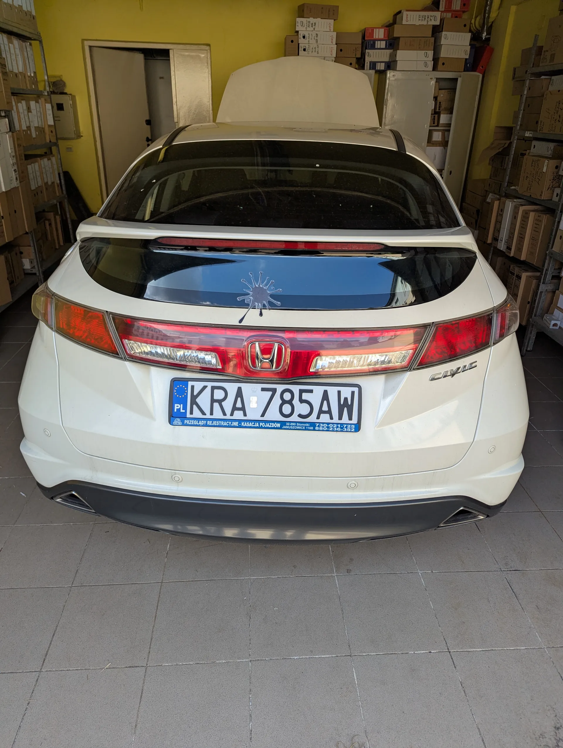 Biały samochód Honda Civic widziany od tyłu, zaparkowany w pomieszczeniu z żółtymi ścianami. Widoczna tablica rejestracyjna PL KRA 785AW z niebieskim pasem zawierającym napisy: PRZEGLĄDY REJESTRACYJNE - KASACJA POJAZDÓW oraz numery telefonów. Na tylnej szybie naklejka w kształcie plamy. W tle regały z kartonami i otwarte drzwi. Podłoga wyłożona szarymi płytkami.