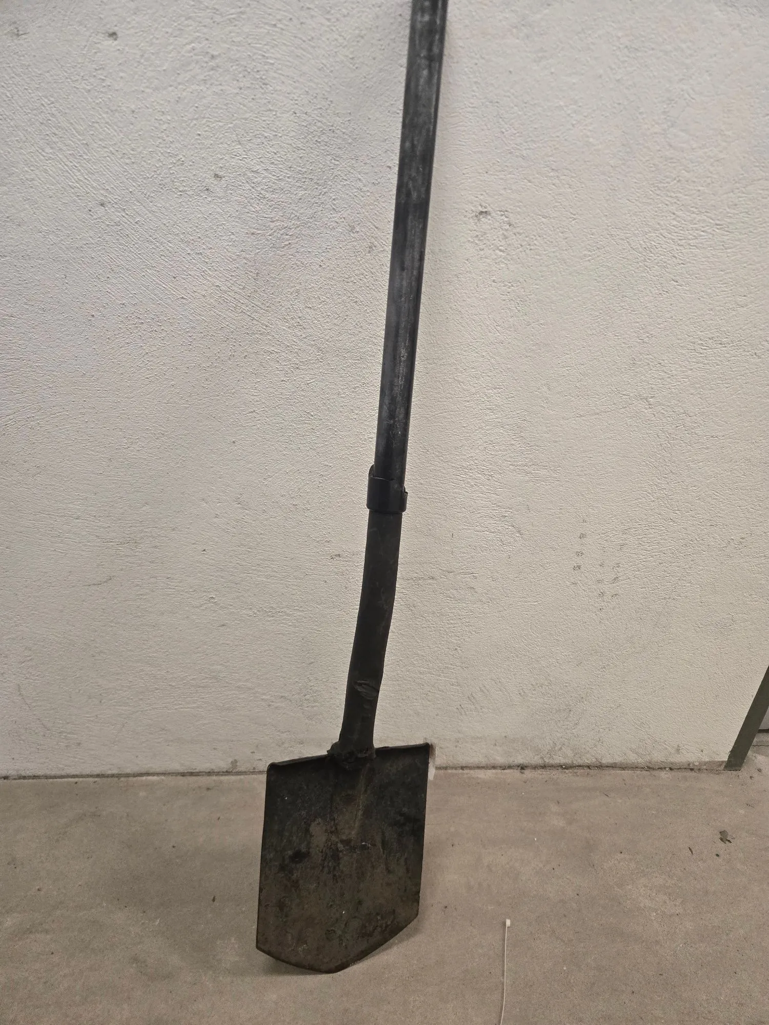 Zdjęcie przedstawiające łopatę opartą o białą, chropowatą ścianę. Łopata ma ciemny, metalowy trzonek i brudną, metalową głowicę, która styka się z szarym, betonowym podłożem.