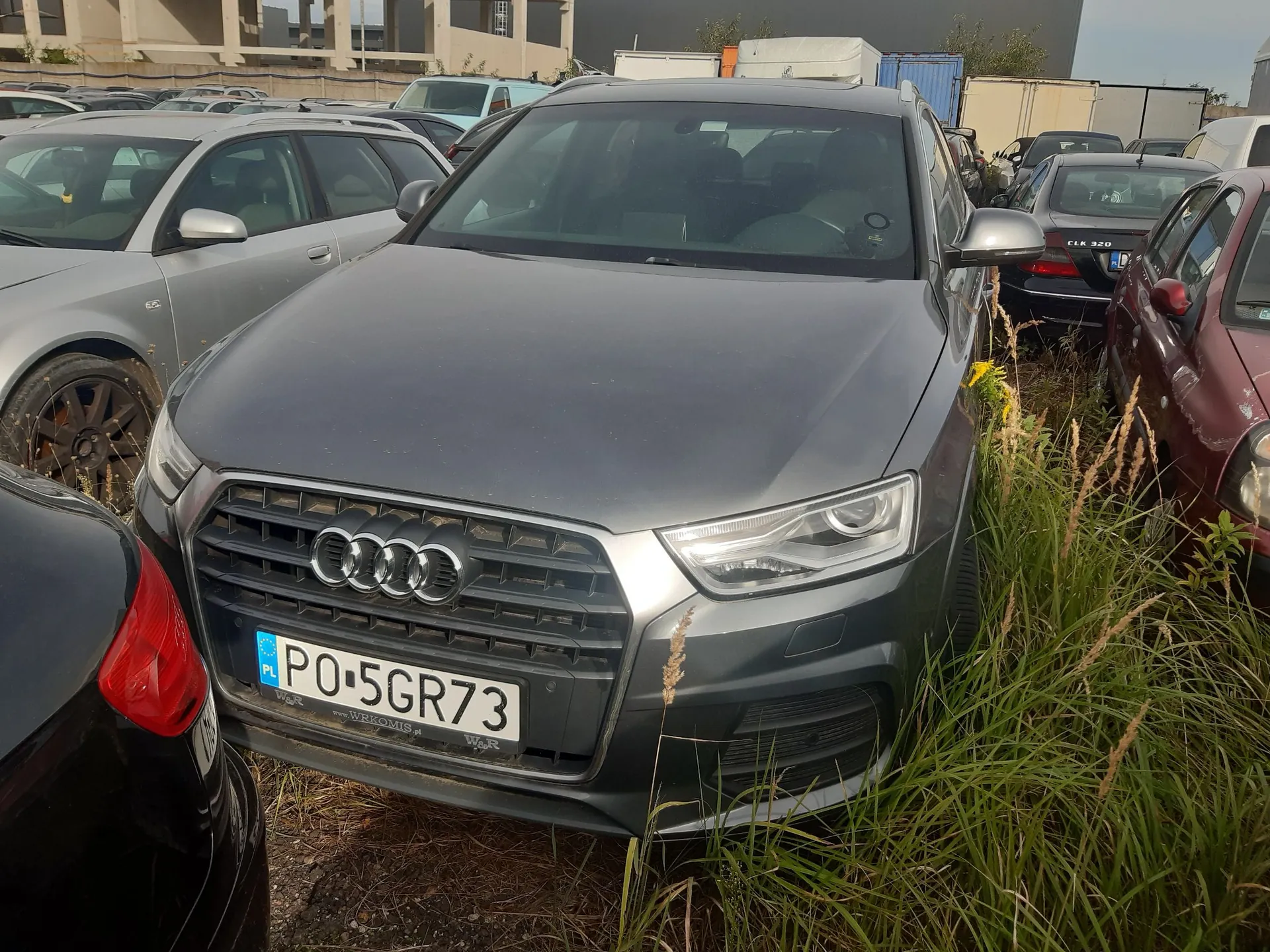 Szary samochód marki Audi zaparkowany na trawiastym terenie obok innych pojazdów. Na przodzie auta widoczna jest charakterystyczna atrapa chłodnicy z czterema pierścieniami oraz polska tablica rejestracyjna o numerze PO 5GR73. W tle znajdują się inne samochody, białe kontenery oraz budynki przemysłowe.