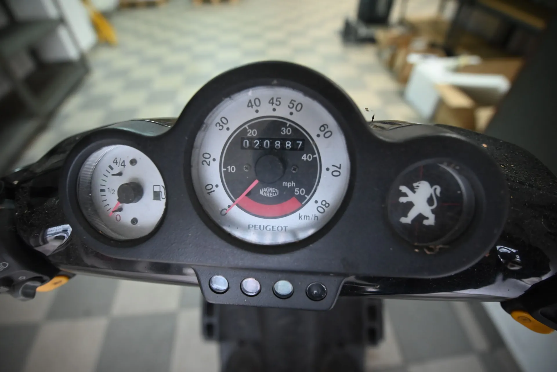 Zbliżenie na panel instrumentów skutera marki Peugeot. W centrum znajduje się okrągły prędkościomierz z podziałką w mph i km/h, z licznikiem przebiegu wskazującym wartość 020887 oraz napisem MAGNETI MARELLI. Po lewej stronie znajduje się mniejszy wskaźnik poziomu paliwa. Po prawej stronie widnieje logo marki Peugeot w formie lwa. Na dole panelu znajduje się napis PEUGEOT oraz cztery małe kontrolki.