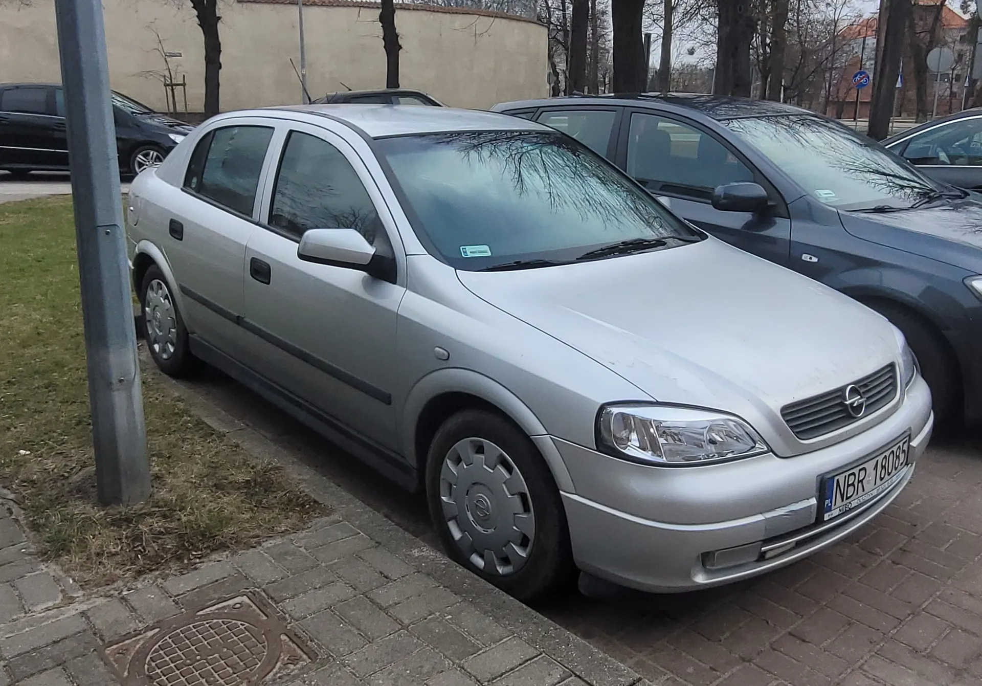Srebrny samochód marki Opel zaparkowany na brukowanej powierzchni. Widoczna tablica rejestracyjna NBR 18085 z kodem PL oraz niebieski naklejka na przedniej szybie. Po lewej stronie metalowy słup, trawa i kanałowa pokrywa. W tle inne zaparkowane pojazdy (ciemnoszary i czarny), beżowy budynek, drzewa bez liści oraz znaki drogowe.