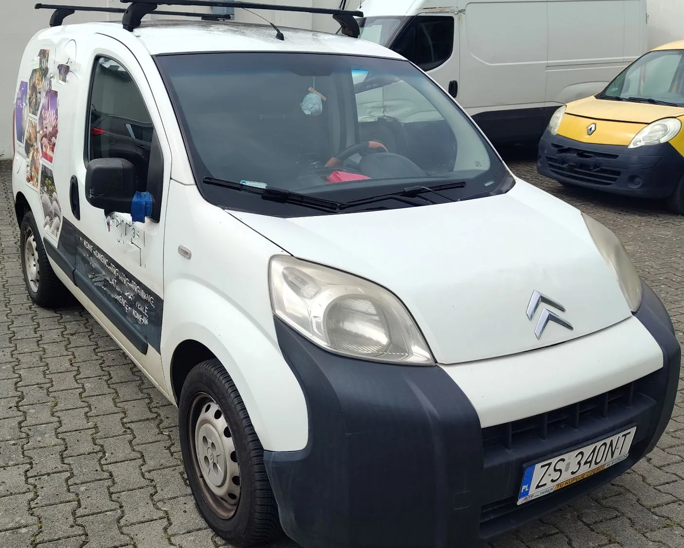 Biały samochód dostawczy marki Citroën zaparkowany na kostce brukowej. Pojazd posiada czarny zderzak, relingi dachowe oraz oklejenie reklamowe na boku z obrazkami i tekstem. Tablica rejestracyjna samochodu to ZS 340NT.