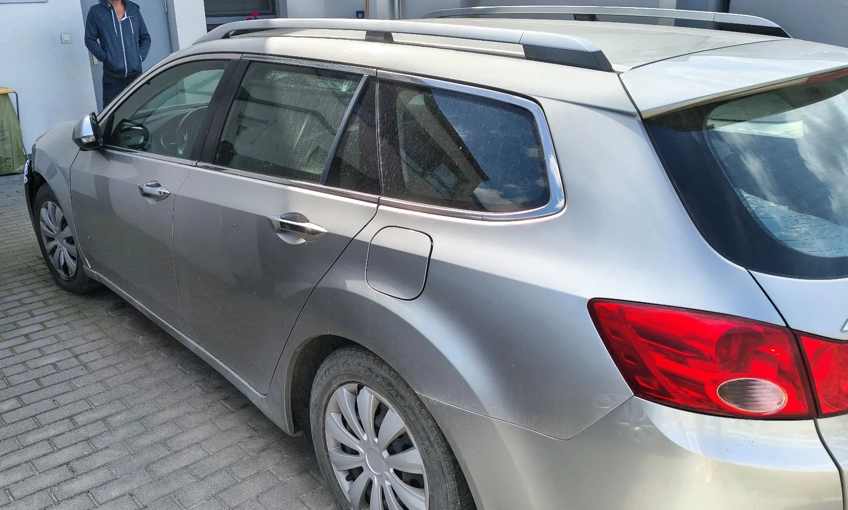 Srebrny samochód typu kombi zaparkowany na kostce brukowej. Zdjęcie przedstawia tylną i boczną część pojazdu, w tym tylne koło z felgą aluminiową, tylną lampę oraz relingi dachowe. W tle widoczny jest fragment budynku oraz osoba w niebieskiej kurtce.