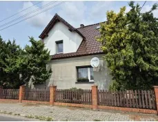 Dom jednorodzinny z białą górą i szarą częścią dolną, czerwoną dachówką, oknem na piętrze i parterze, satelitą na ścianie. Przed domem drewniane ogrodzenie z ceglanymi filarami, drzewa po obu stronach, brukowana droga i częściowo zachmurzone niebo.