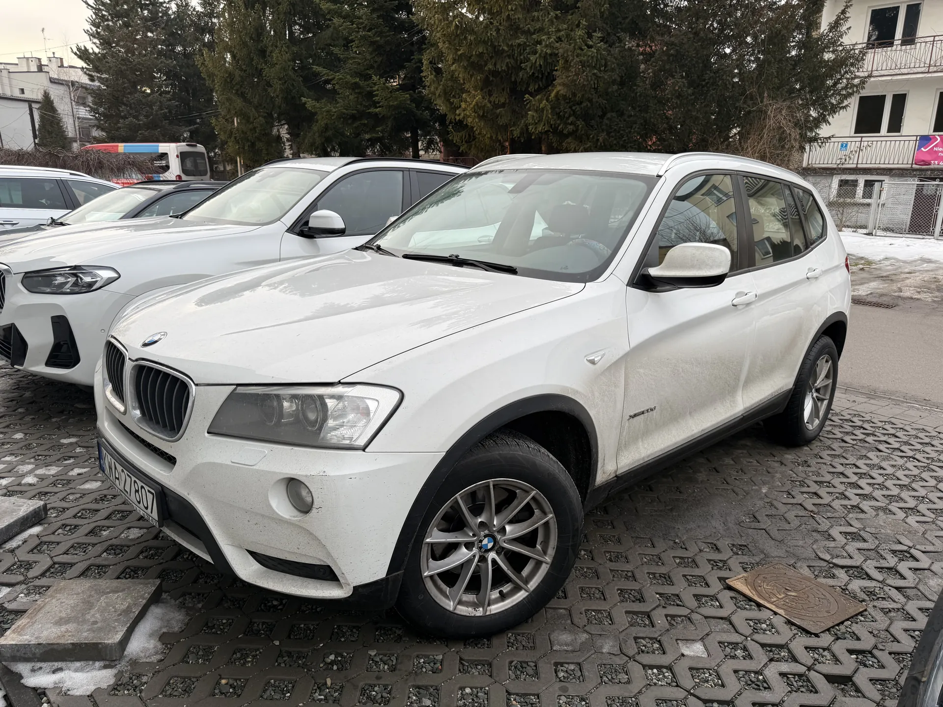 Biały samochód SUV marki BMW zaparkowany na brukowanej powierzchni z widoczną tablicą rejestracyjną WA 27807. W tle inne białe samochody, drzewa, budynki oraz resztki śniegu na ziemi.