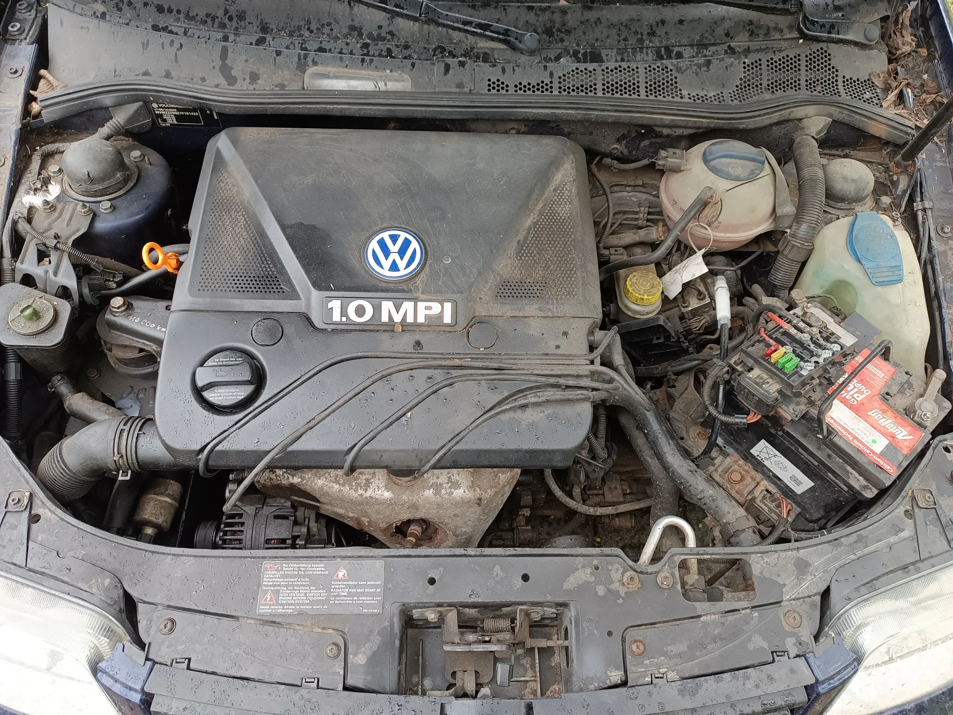 Silnik samochodu marki Volkswagen z pokrywą oznaczoną napisem „1.0 MPI” i logo VW. Widoczne są różne elementy komory silnikowej: zbiornik chłodniczy z niebieską pokrywką, zbiornik płynu hamulcowego z niebieską pokrywką, bezpieczniki w kolorach czerwonym, żółtym i zielonym, bateria z czerwoną etykietą oraz przewody i rury. Komora silnikowa jest zabrudzona, z widocznymi śladami brudu i liśćmi wokół krawędzi. Klapa silnika jest otwarta, a reflektory przednie są częściowo widoczne.